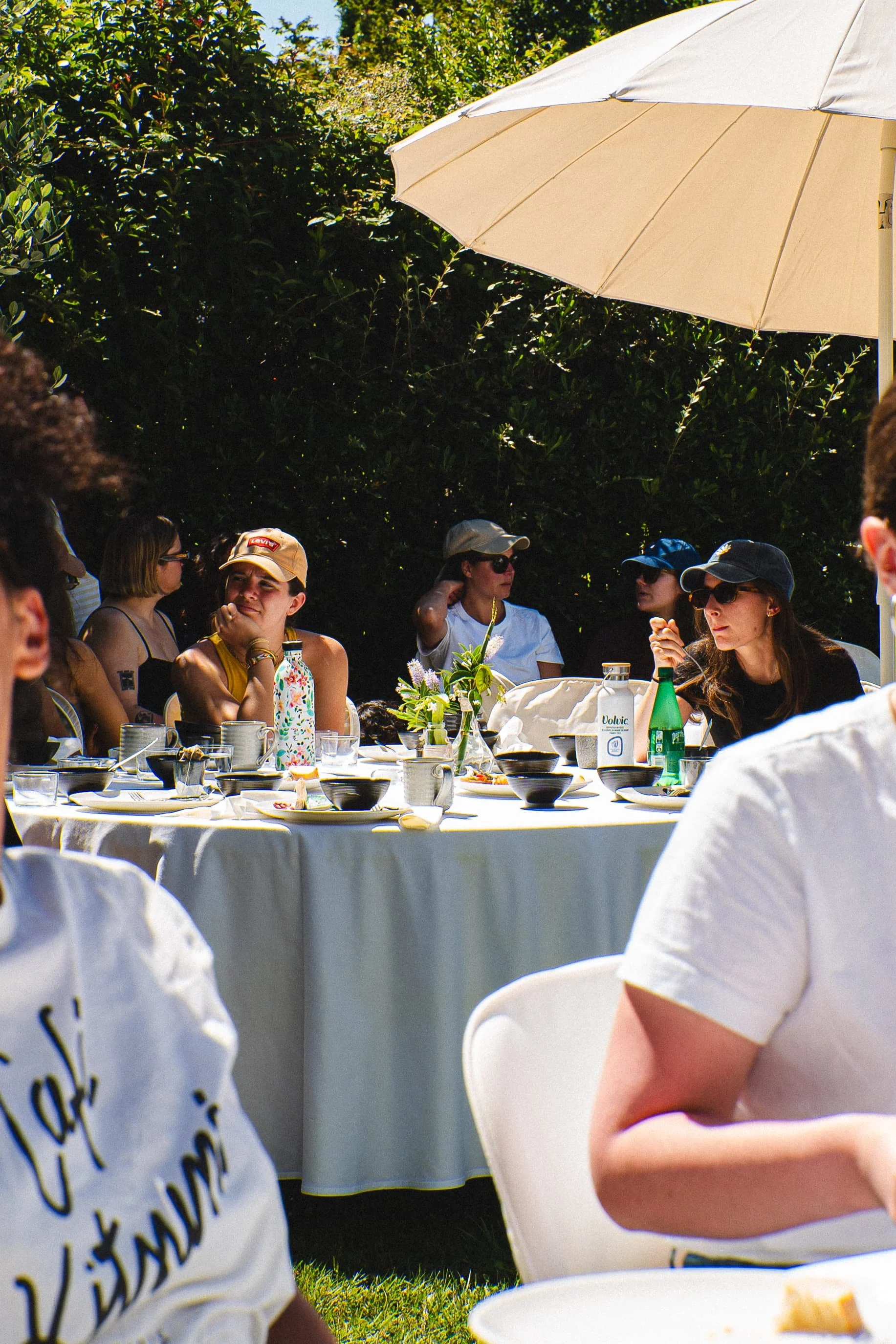 Groupe de personnes assises autour d'une table lors d'un repas en plein air, avec un parasol blanc en arrière-plan, dans un environnement verdoyant