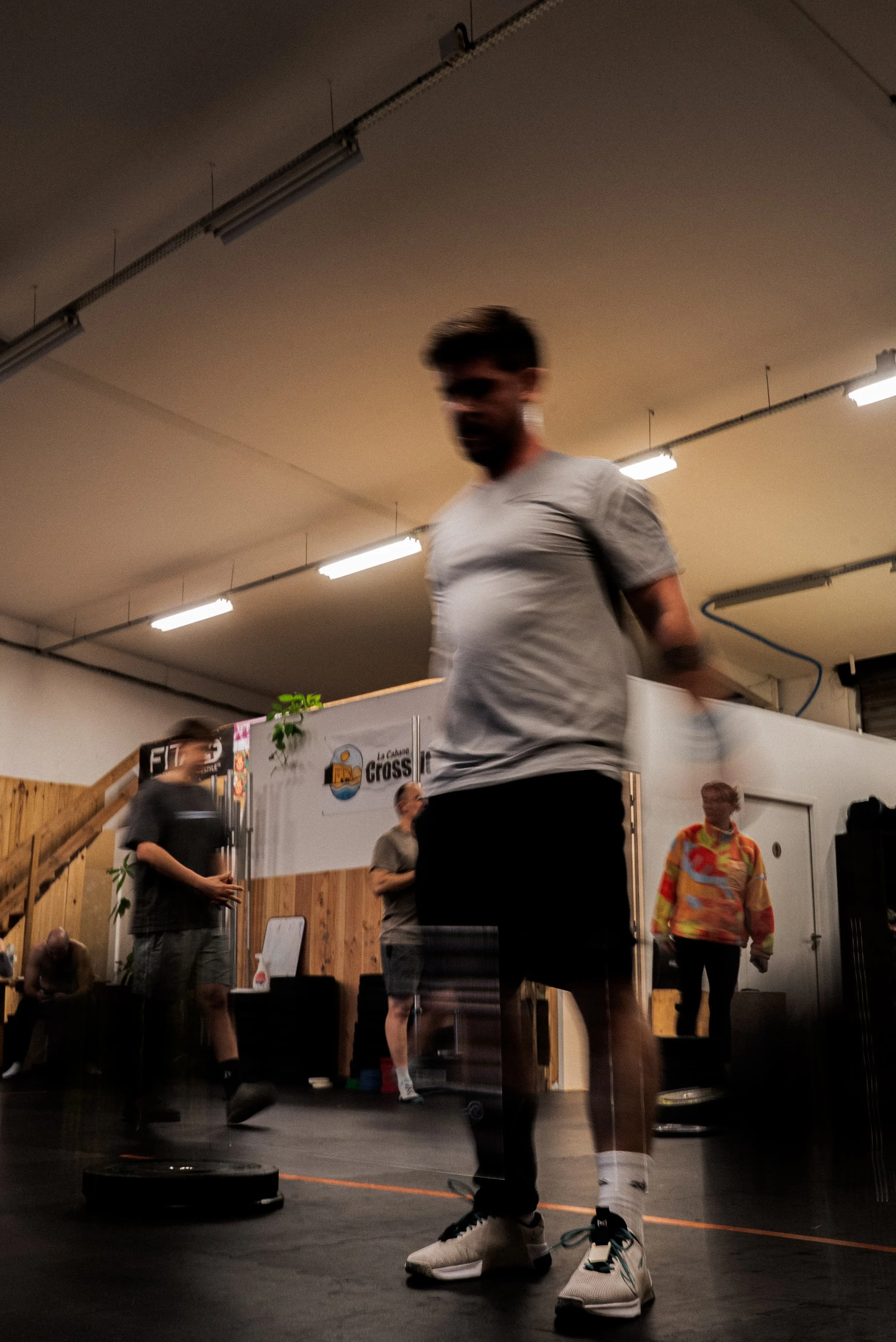Un homme en t-shirt gris et pantalons noirs effectue un exercice de musculation, probablement du saut. Il porte des baskets blanches et se trouve dans un espace intérieur avec un sol en bois foncé, des murs en bois clair, et un plafond avec des lumin