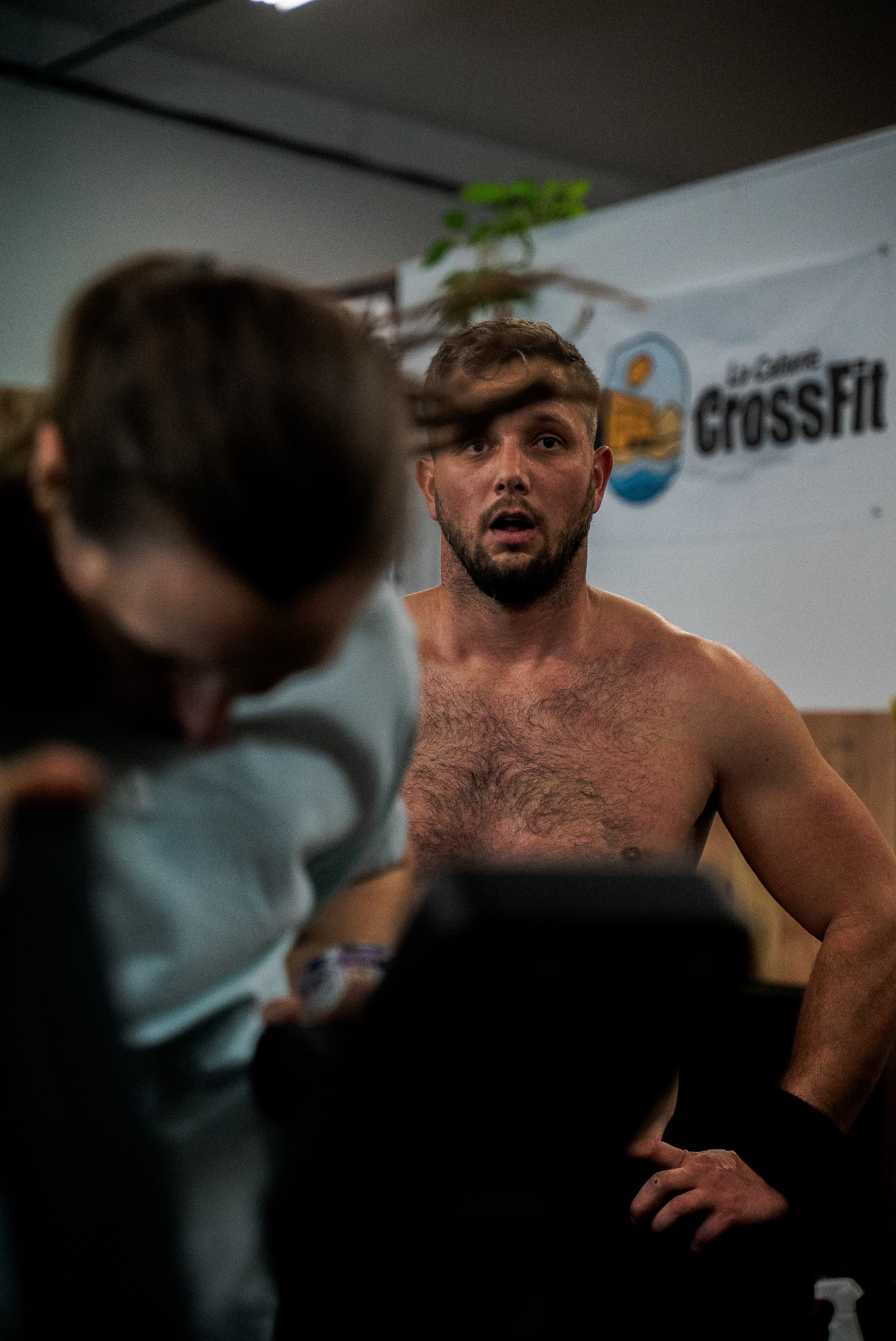 Un homme torse nu avec une barbe, en train de parler ou écouter, dans un environnement intérieur avec un logo 'CrossFit' en arrière-plan.