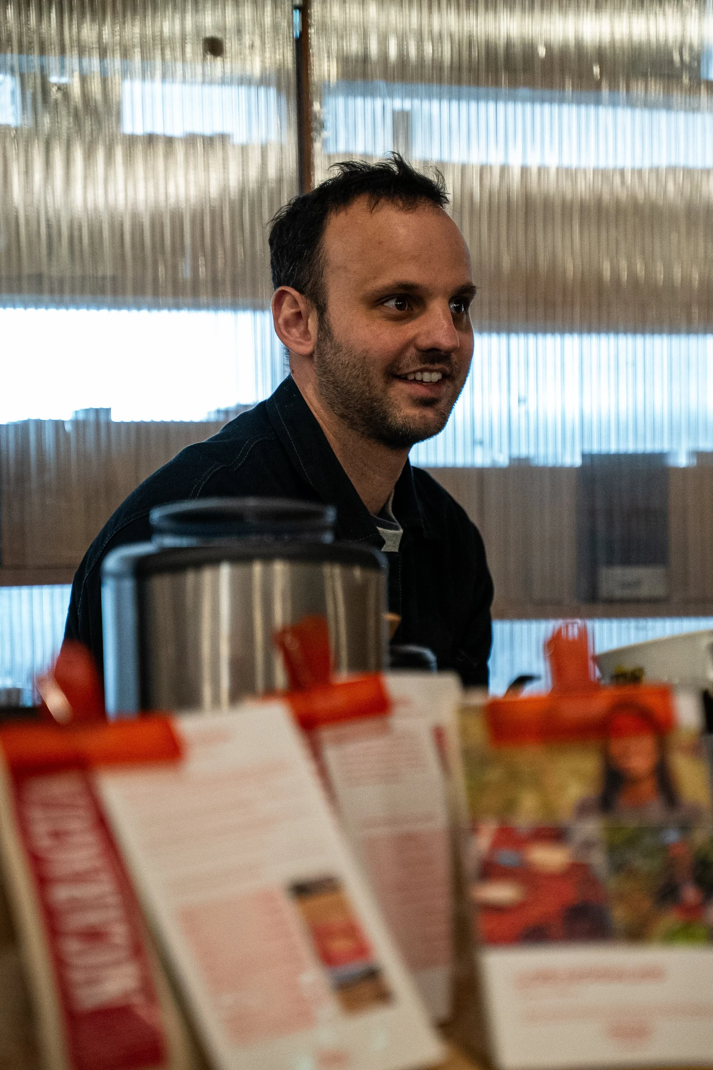 Un homme souriant dans un intérieur, avec des rayures de lumière à l'arrière-plan, assis derrière une table avec des produits ou des brochures en avant-plan.