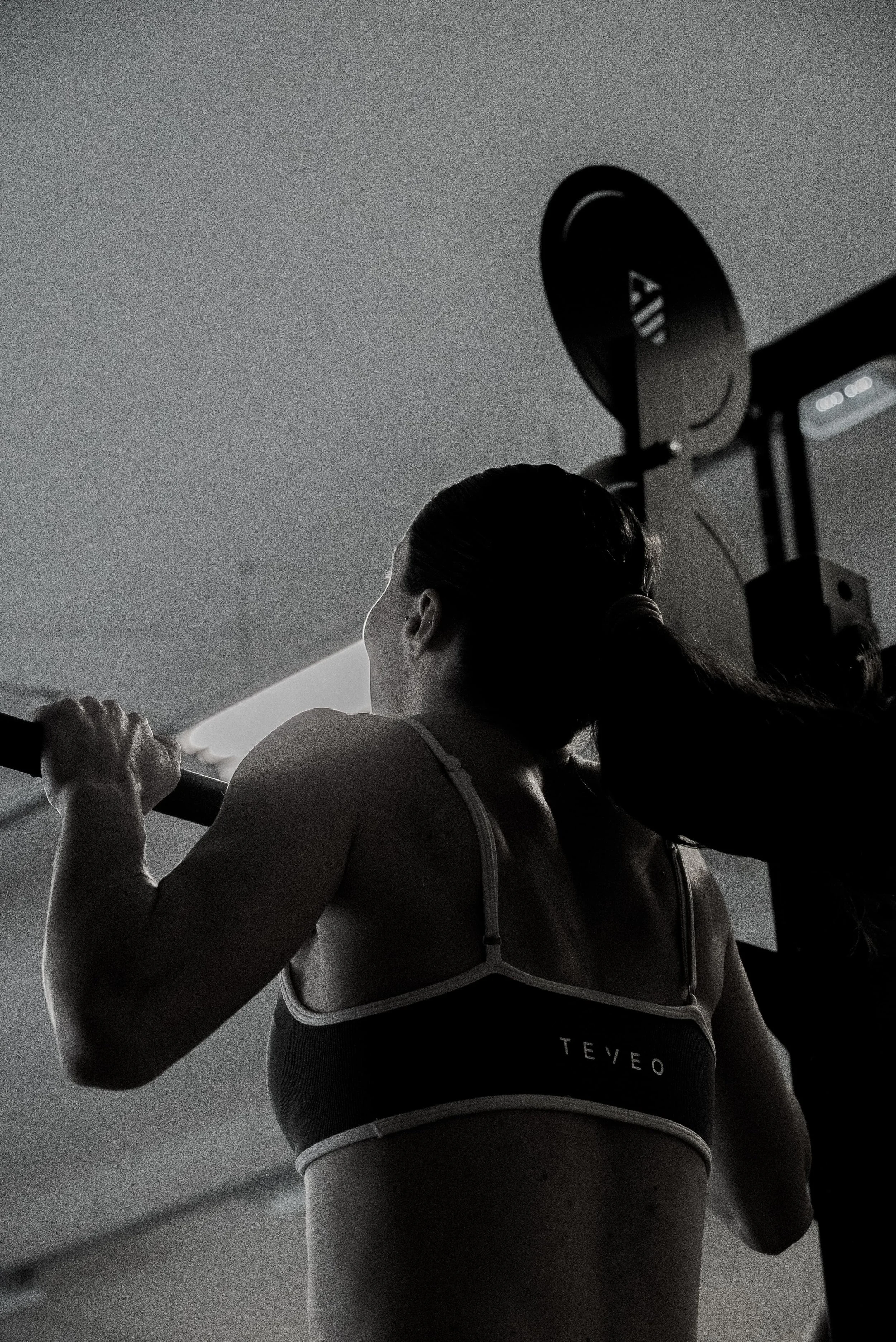Une femme en tenue de sport, vue de dos, faisant de la musculation avec une barre dans une salle de sport. L'image est en noir et blanc.