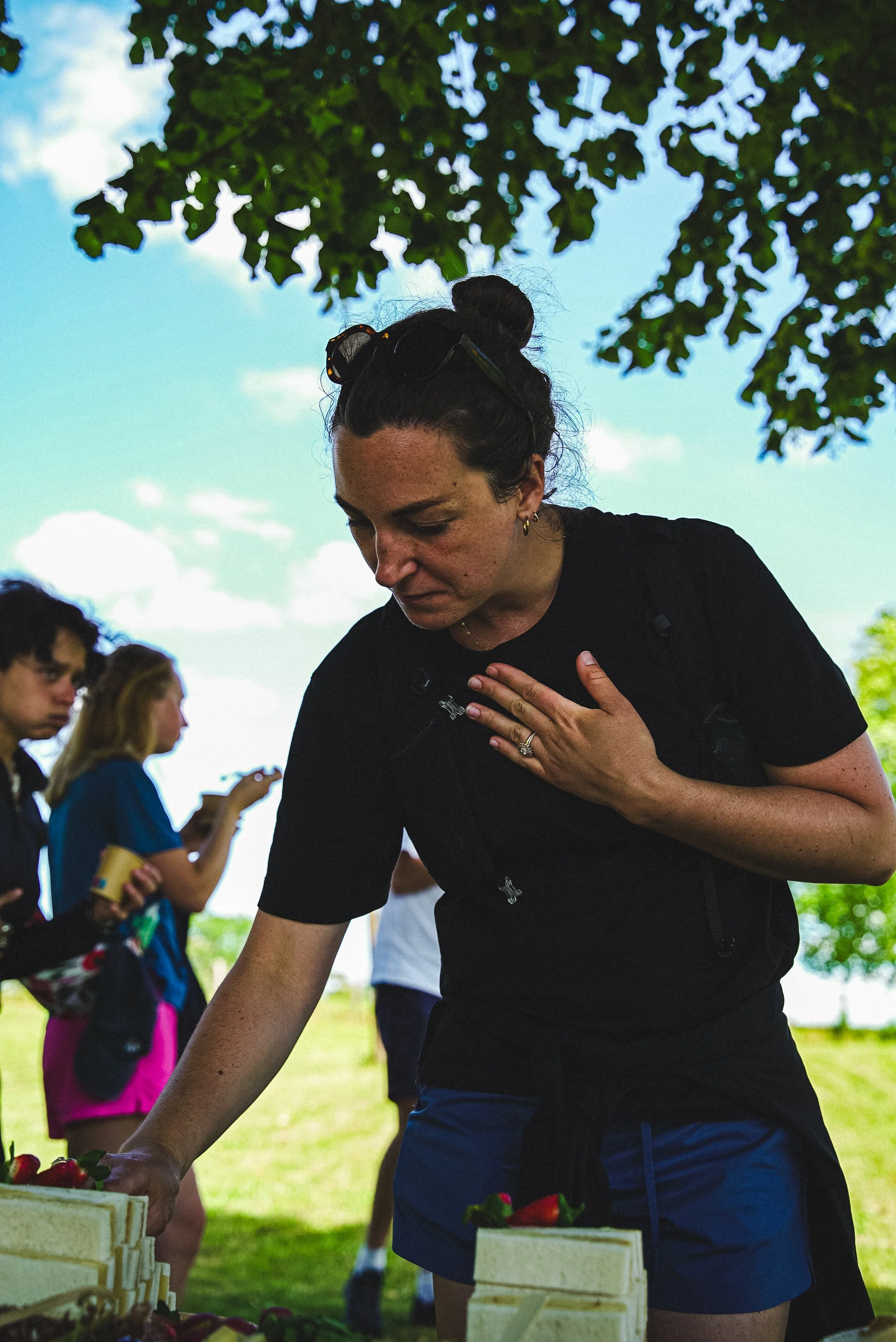 Une femme en t-shirt noir et short bleu, avec des lunettes sur la tête, penchée sur une table en plein air, semble préparer ou servir des aliments. Elle a une main sur la poitrine et une autre sur la table. En arrière-plan, des personnes, notamment d