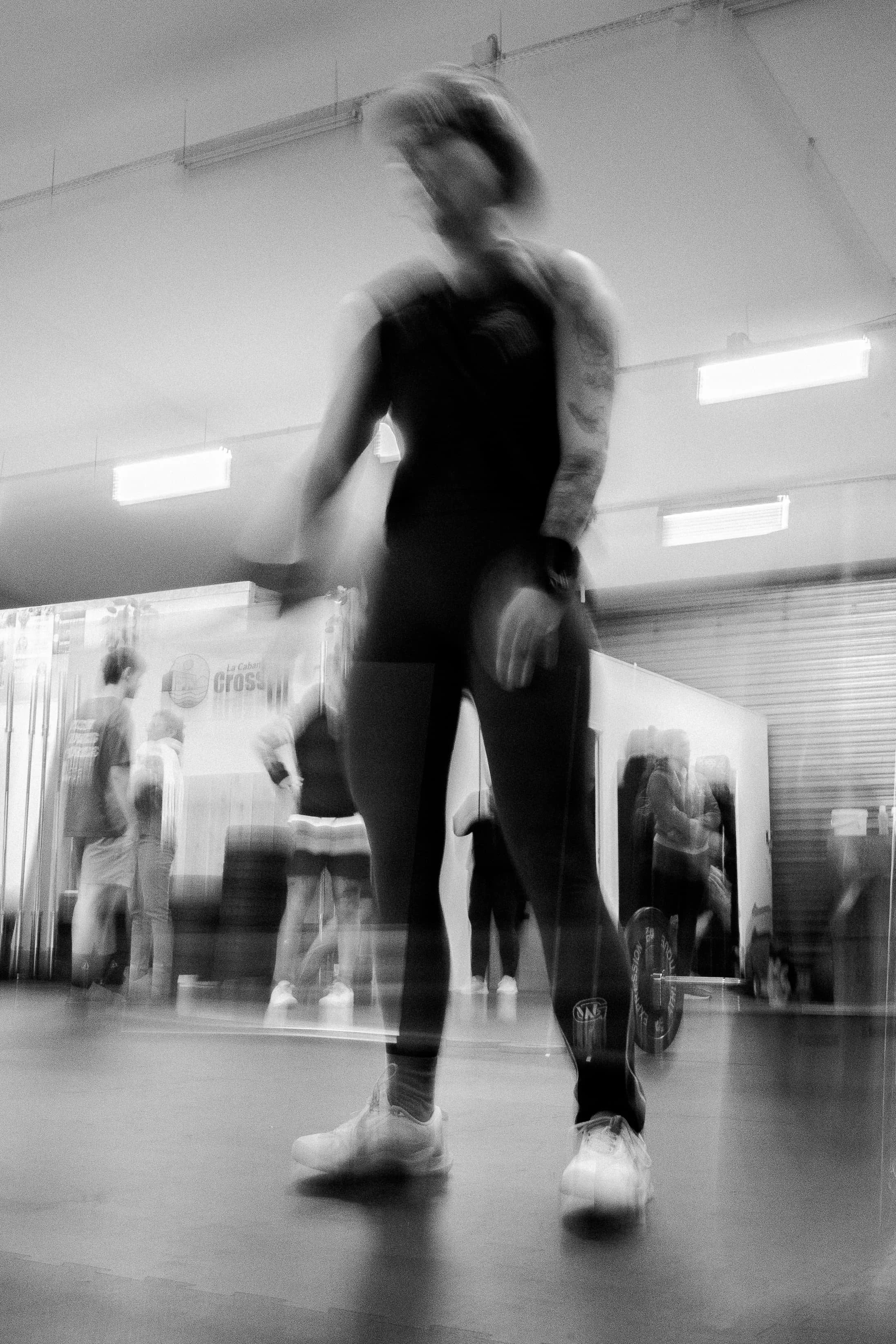 Une personne blonde avec des tatouages, portant un débardeur noir et un masque, en train de danser ou pratiquer une activité physique dans une salle de sport ou de danse, avec un mur miroir derrière eux refletant d'autres personnes.