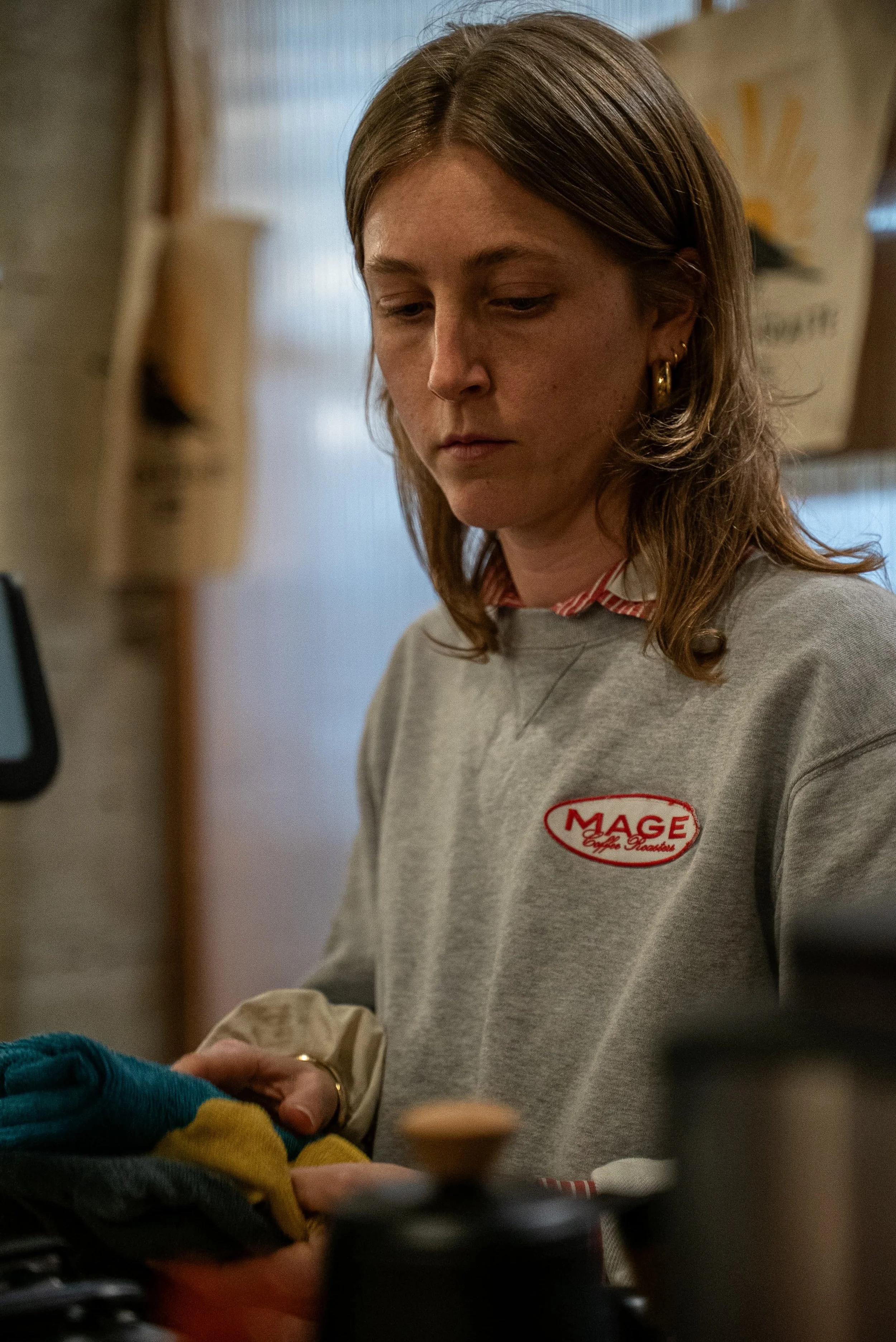 Une femme portant un sweat gris avec un logo « MAGE » en rouge, en train de servir ou de préparer quelque chose dans une cuisine ou un endroit semblable.
