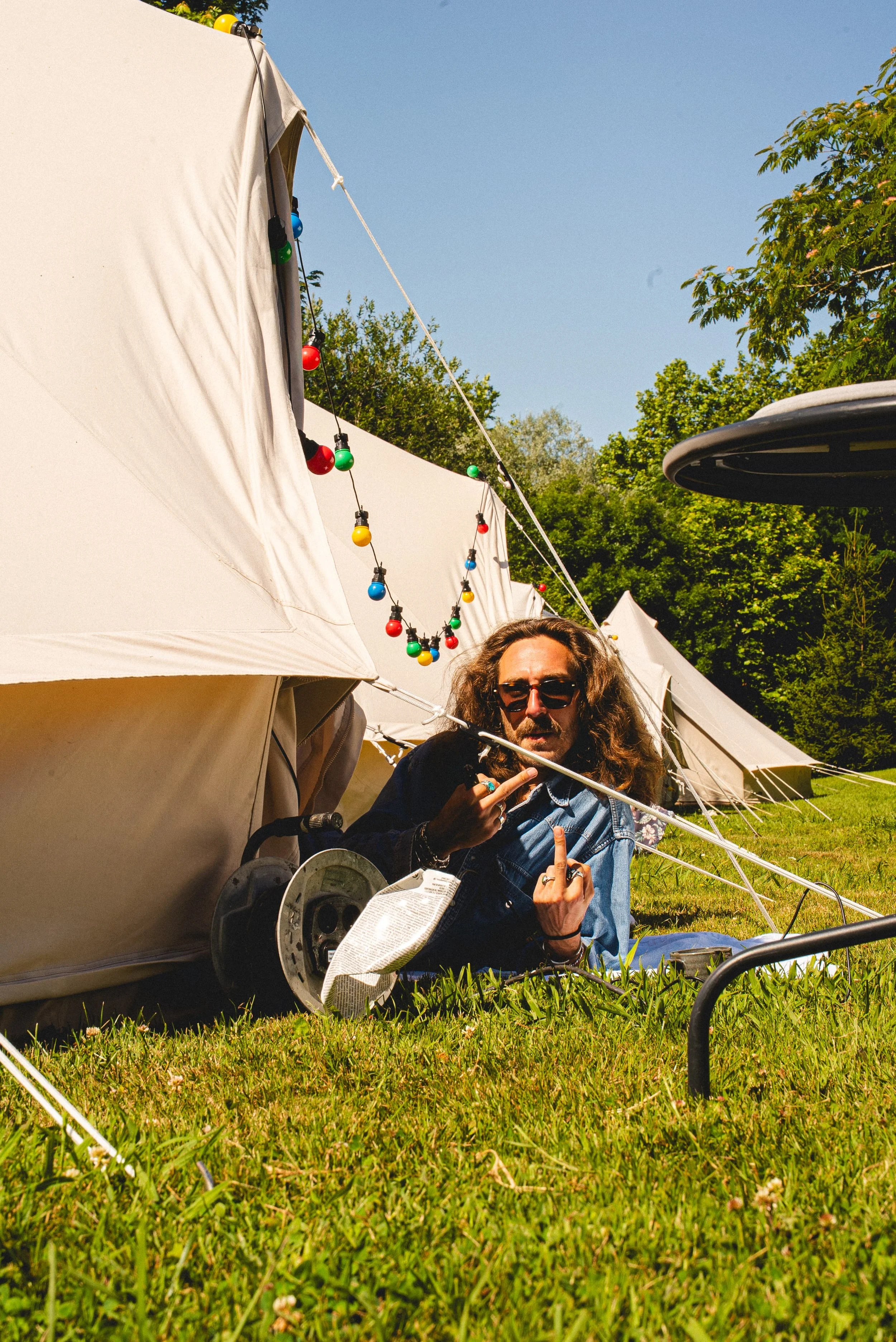 Une personne assise sur l'herbe devant une tente, portant des lunettes de soleil, avec un casque de moto à ses côtés, tenant un ordinateur portable et faisant un geste avec la main. La tente est décorée de guirlandes lumineuses colorées. Il y a d'aut
