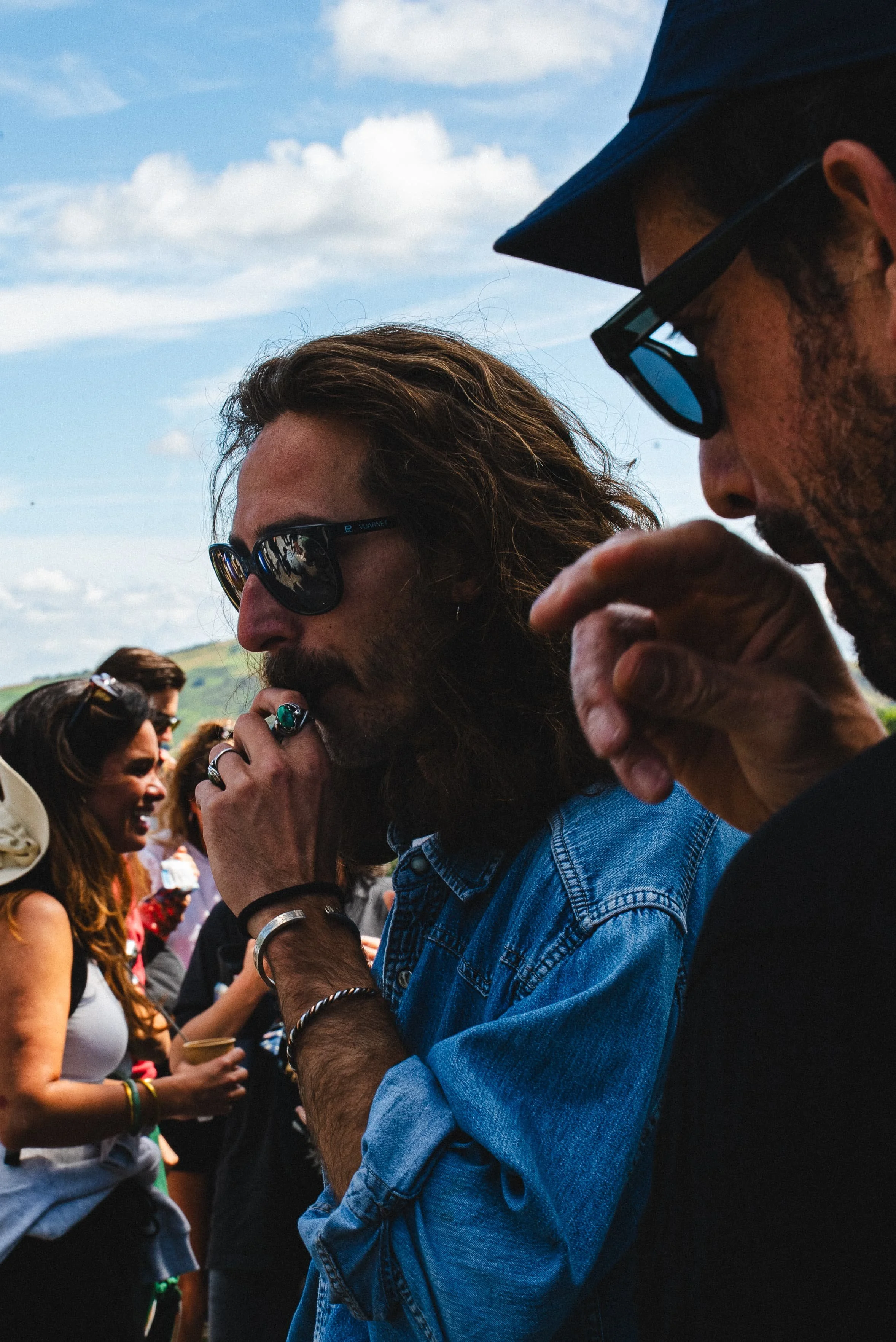 Groupe de jeunes adultes à l'extérieur, certains portent des lunettes de soleil, en train de discuter lors d'un événement en plein air sous un ciel partiellement nuageux.