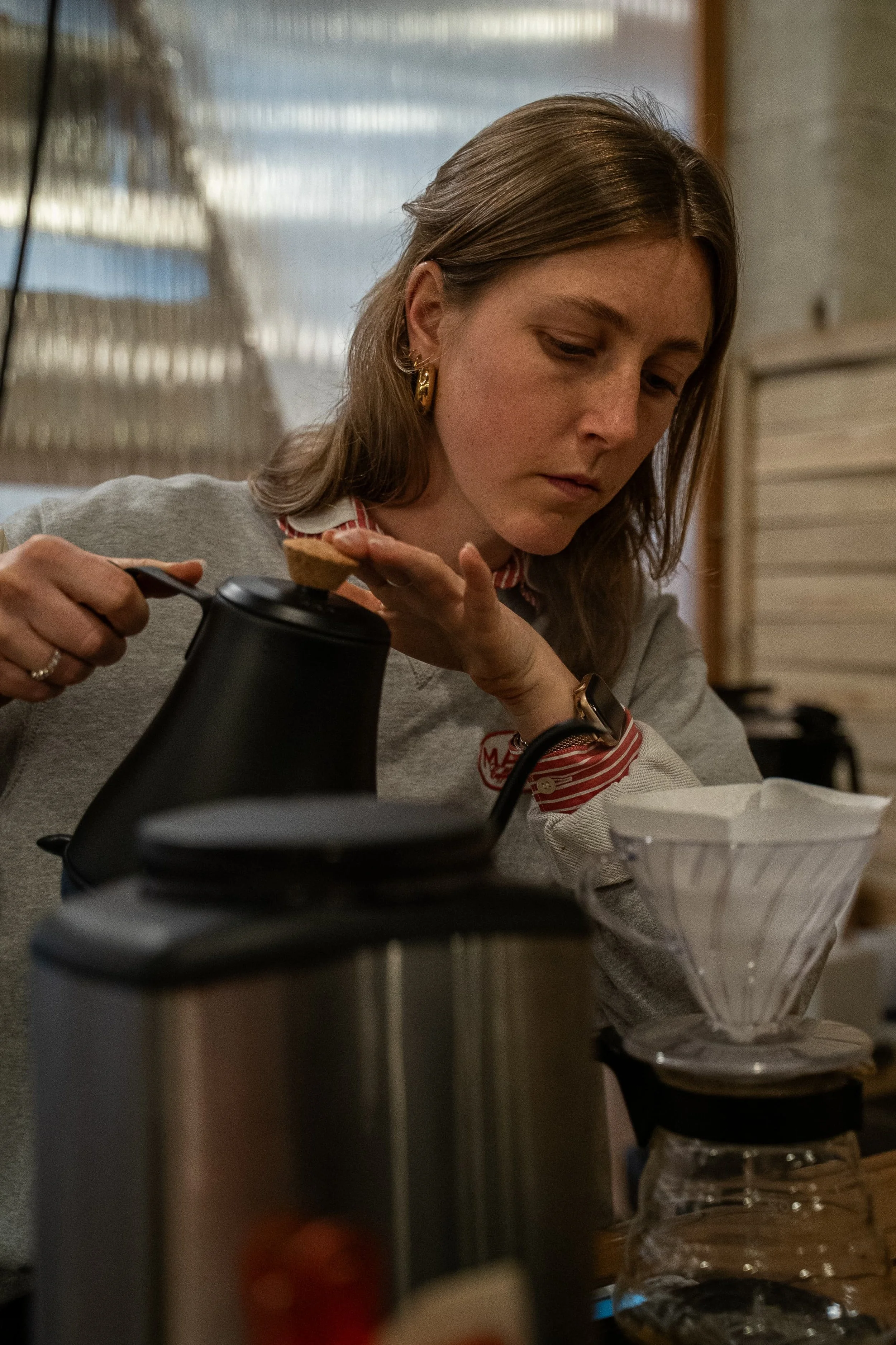 Une femme brune regarde vers le bas devant une machine à café avec une tasse en bas, dans une cuisine. Il y a aussi un entonnoir en plastique avec un contenu pâteux ou poudreux.
