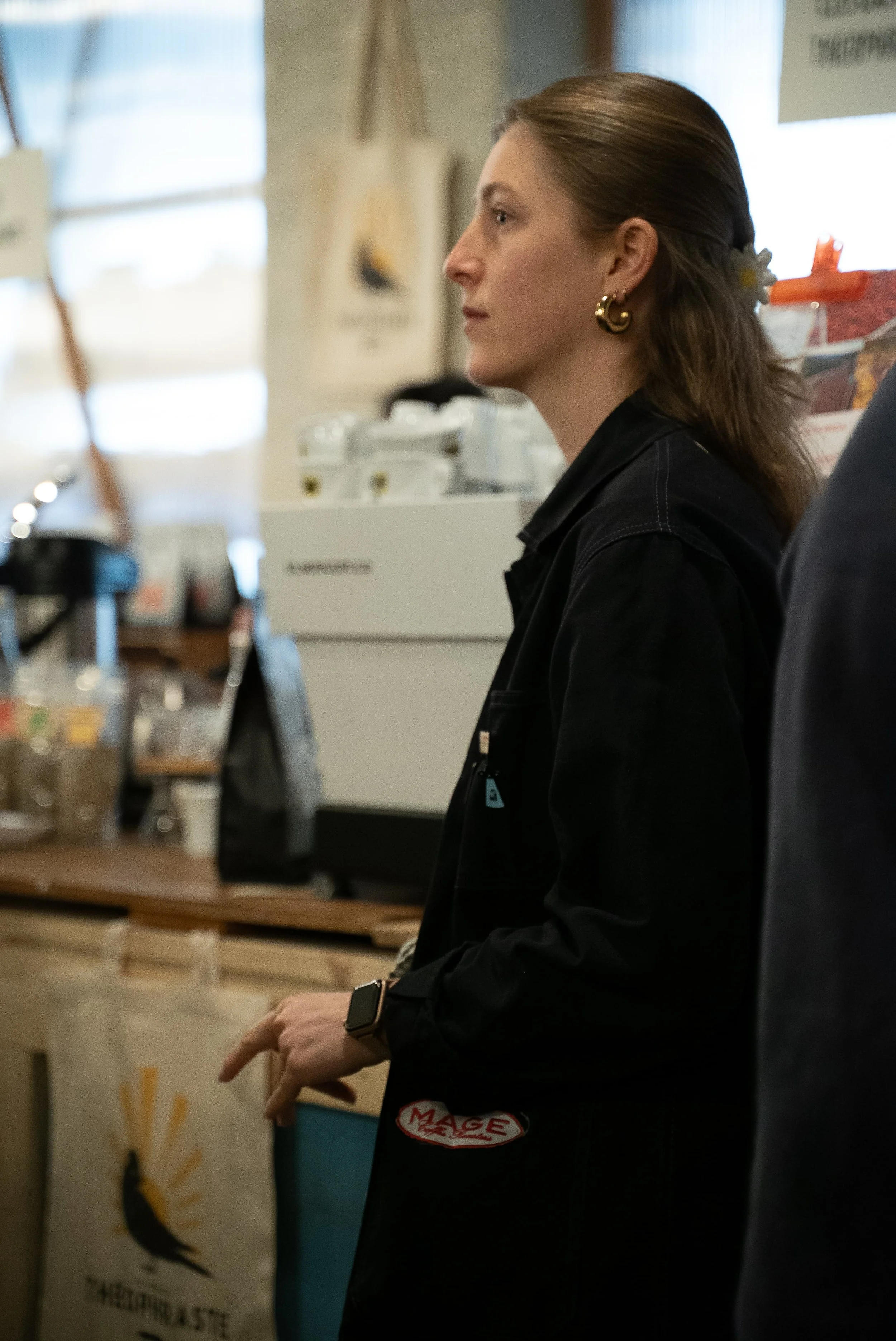 Femme en profil portant un blazer noir, adossée à un comptoir dans un café ou restaurant, avec des affiches et décorations en arrière-plan.
