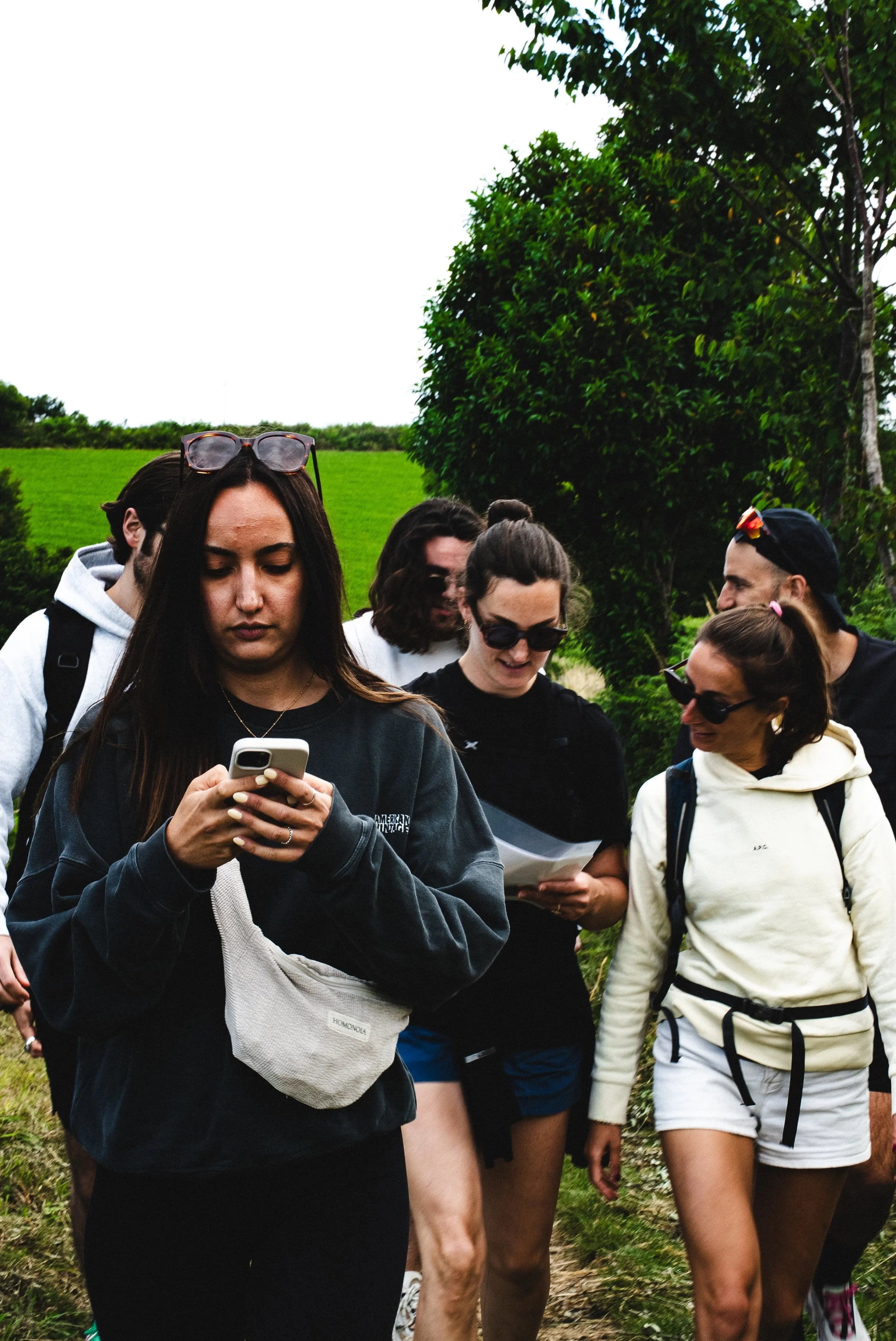 Groupe de jeunes femmes et hommes marchant dans la nature, certaines portant des sacoches ou des sacs à dos, portant des vêtements décontractés, sous un arbre et un ciel nuageux.