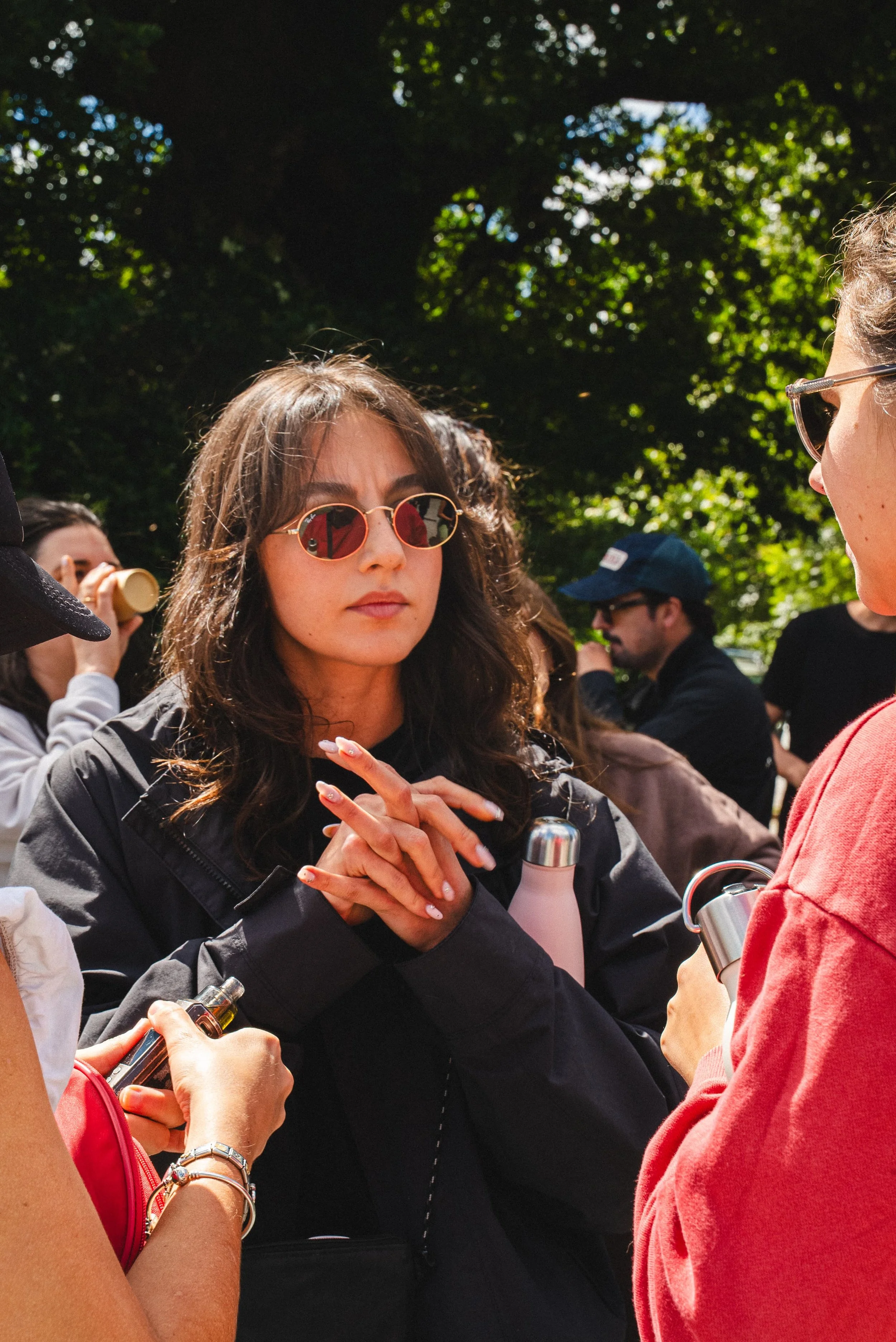 Groupe de personnes discutant à l'extérieur sous un arbre, femmes avec lunettes de soleil, vêtements décontractés, ambiance en plein air.
