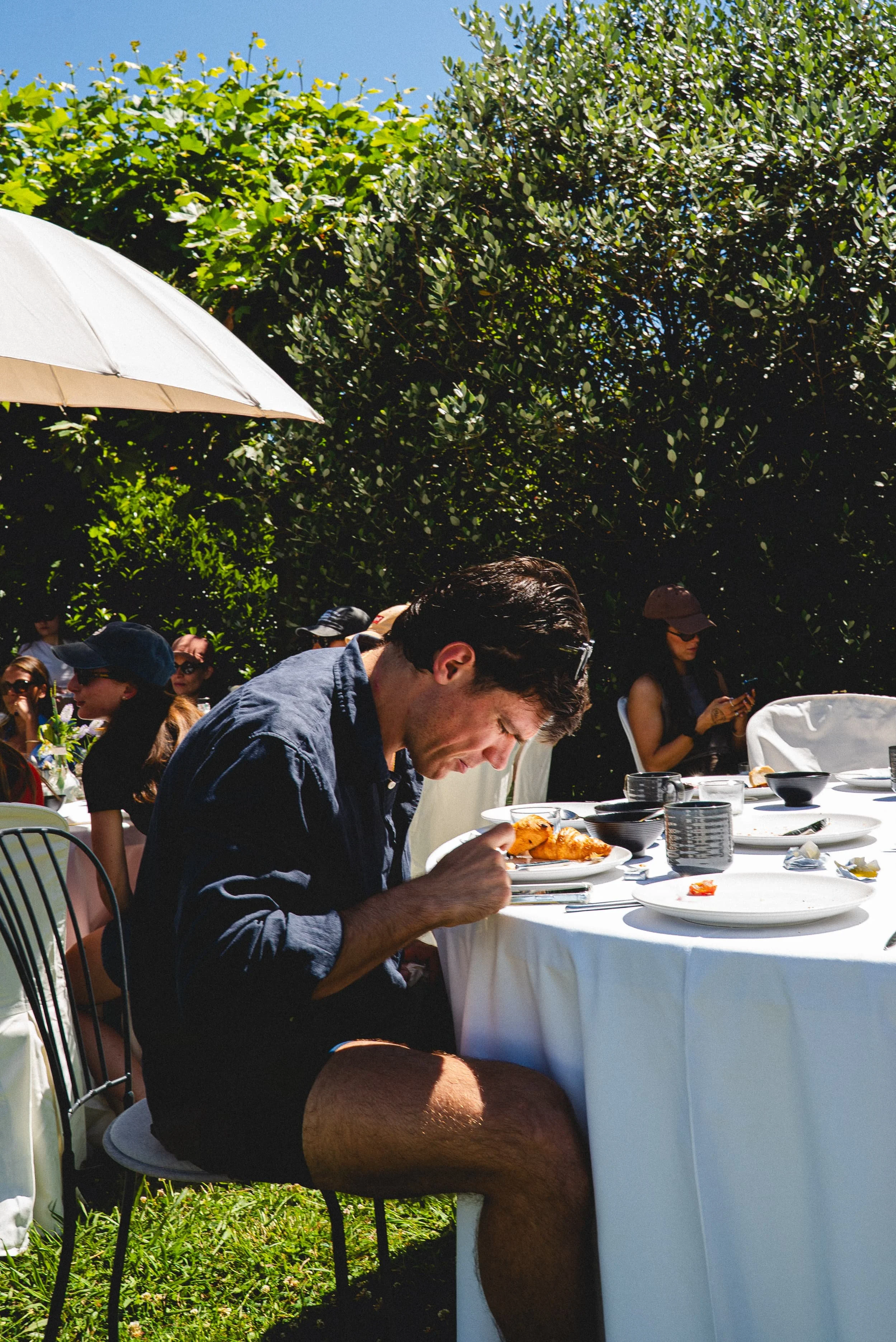 Un homme mange un repas dans un cadre extérieur avec un groupe de personnes assises autour d'une table ronde, entouré de végétation verdoyante et un parasol blanc.