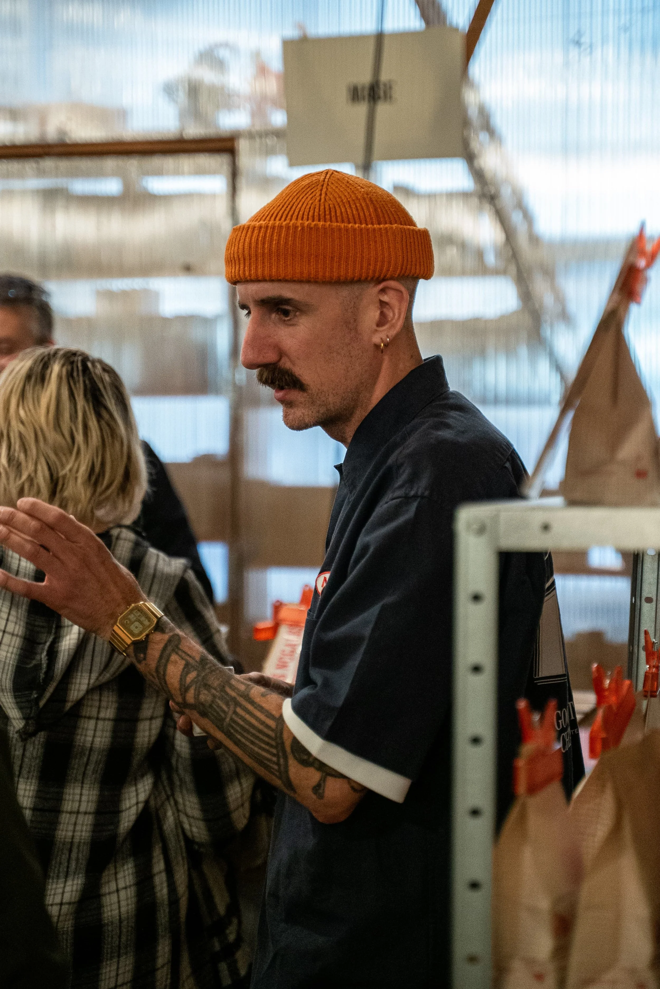 Un homme portant un bonnet orange, une chemise noire et ayant des tatouages visibles sur les bras, échange des mots ou un objet avec une autre personne dans un environnement intérieur.