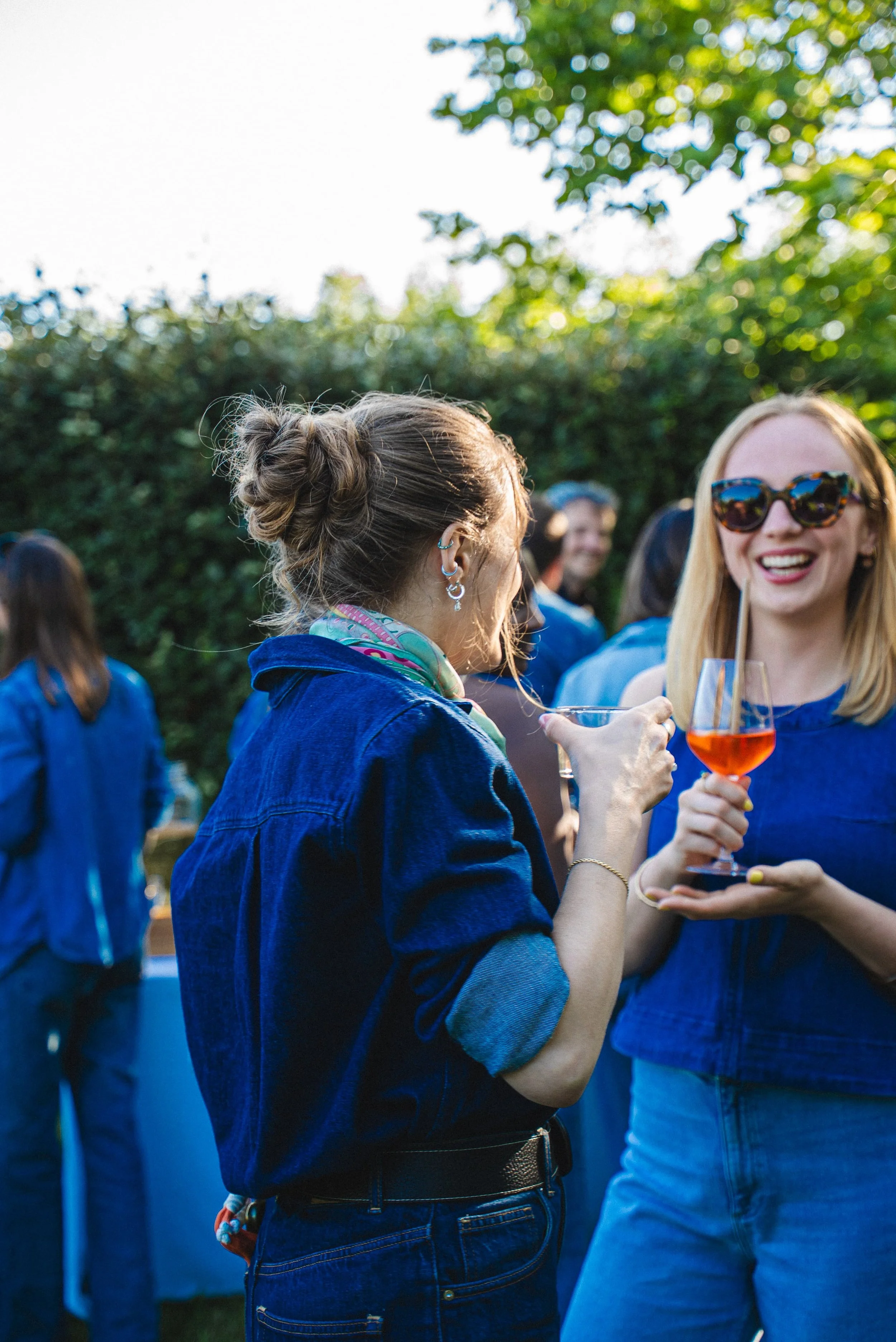 Deux femmes qui sourient, portant des jeans et des vêtements bleus, en train de trinquer avec des verres de vin rosé lors d'un rassemblement en extérieur, entourées d'autres personnes.