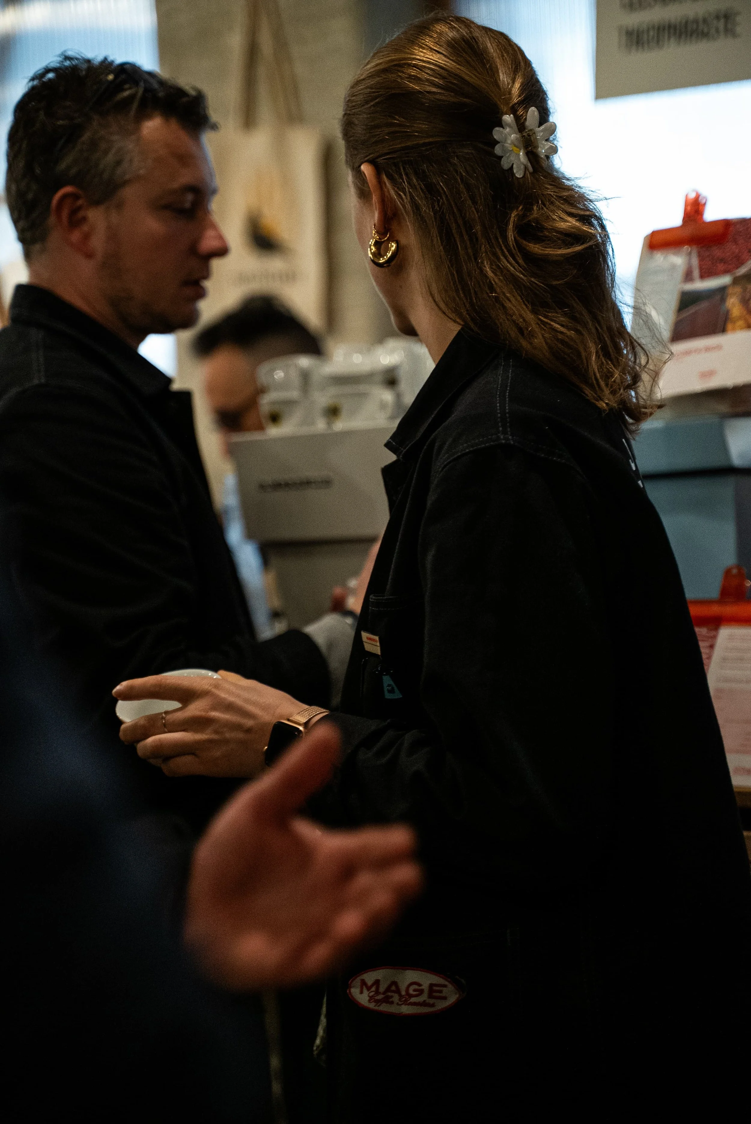 Deux personnes discutent dans un endroit intérieur, apparemment à une caisse ou un comptoir, avec une boîte en arrière-plan. L'une porte une veste noire et une montre, l'autre porte des boucles d'oreilles en forme de cercles et un accessoire de cheve