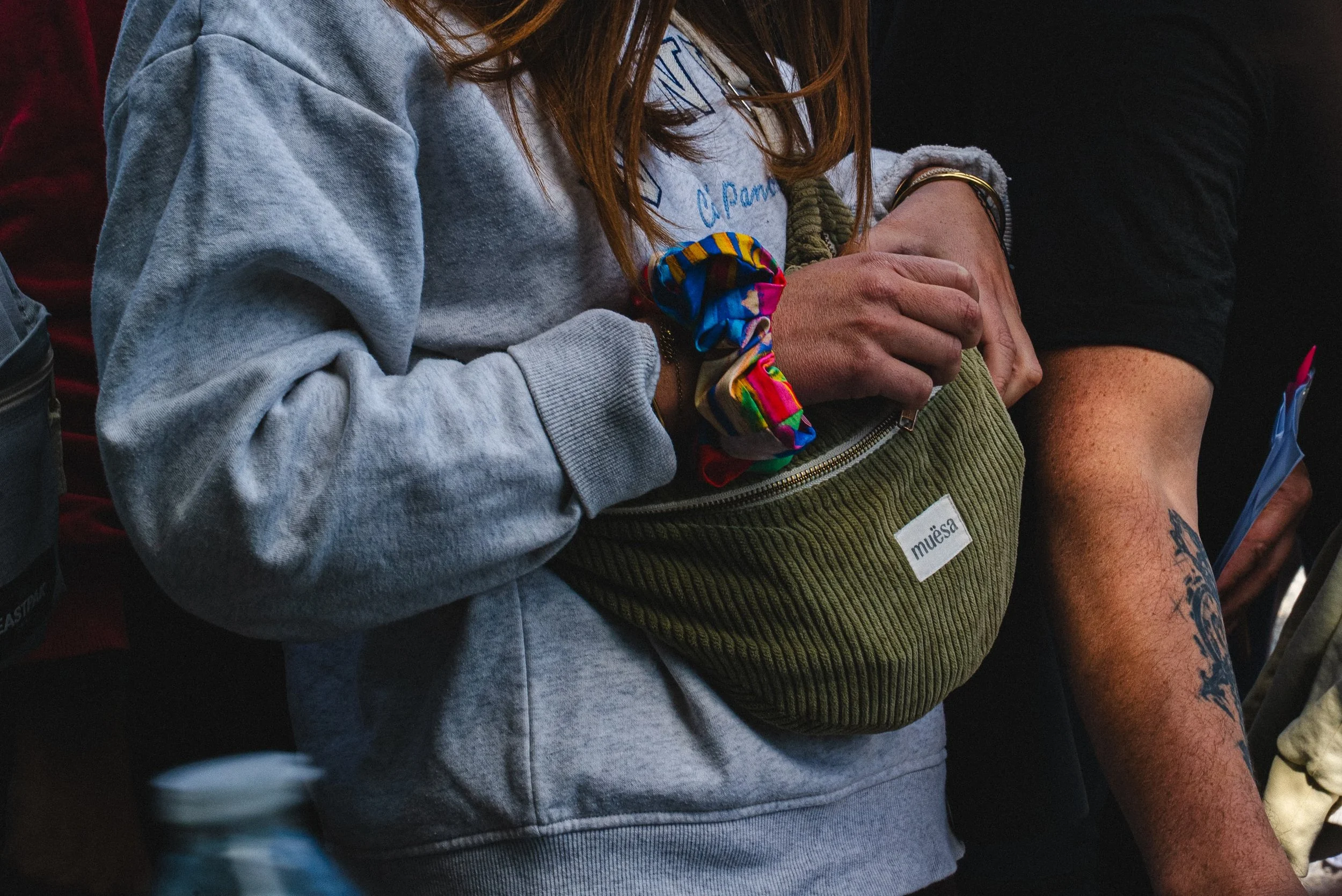 Gros plan sur deux personnes, une portant une veste grise et l'autre une poche vert olive. La personne avec la poche porte un bracelet noir et une bague, et a un tatouage sur le bras. La scène semble être dans un endroit partagé et informel.