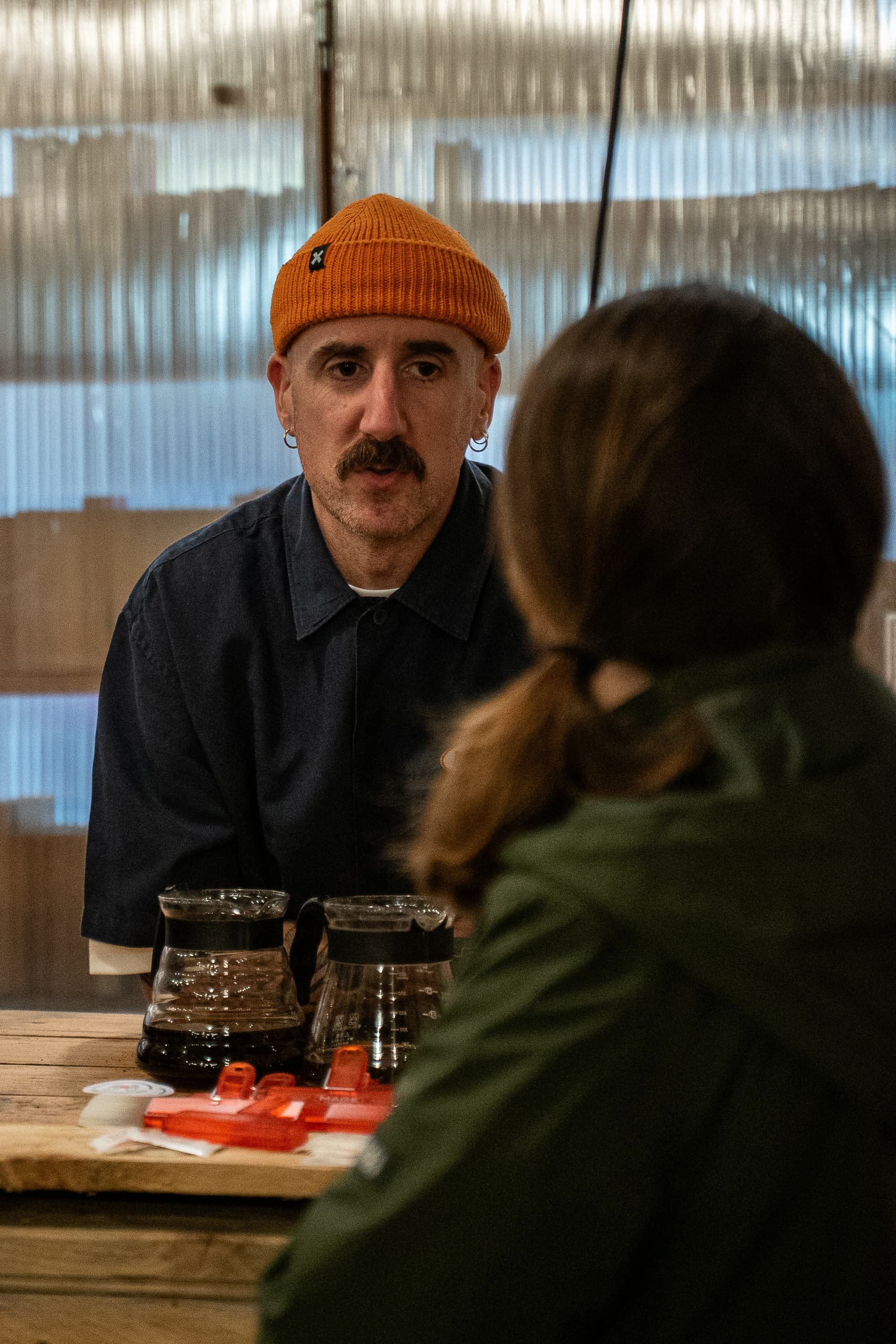 Un homme portant un bonnet orange et une chemise foncée est assis à une table en bois, parlant à une personne dont le visage est flou et partiellement visible. Sur la table, il y a des carafes de café et des objets rouges.