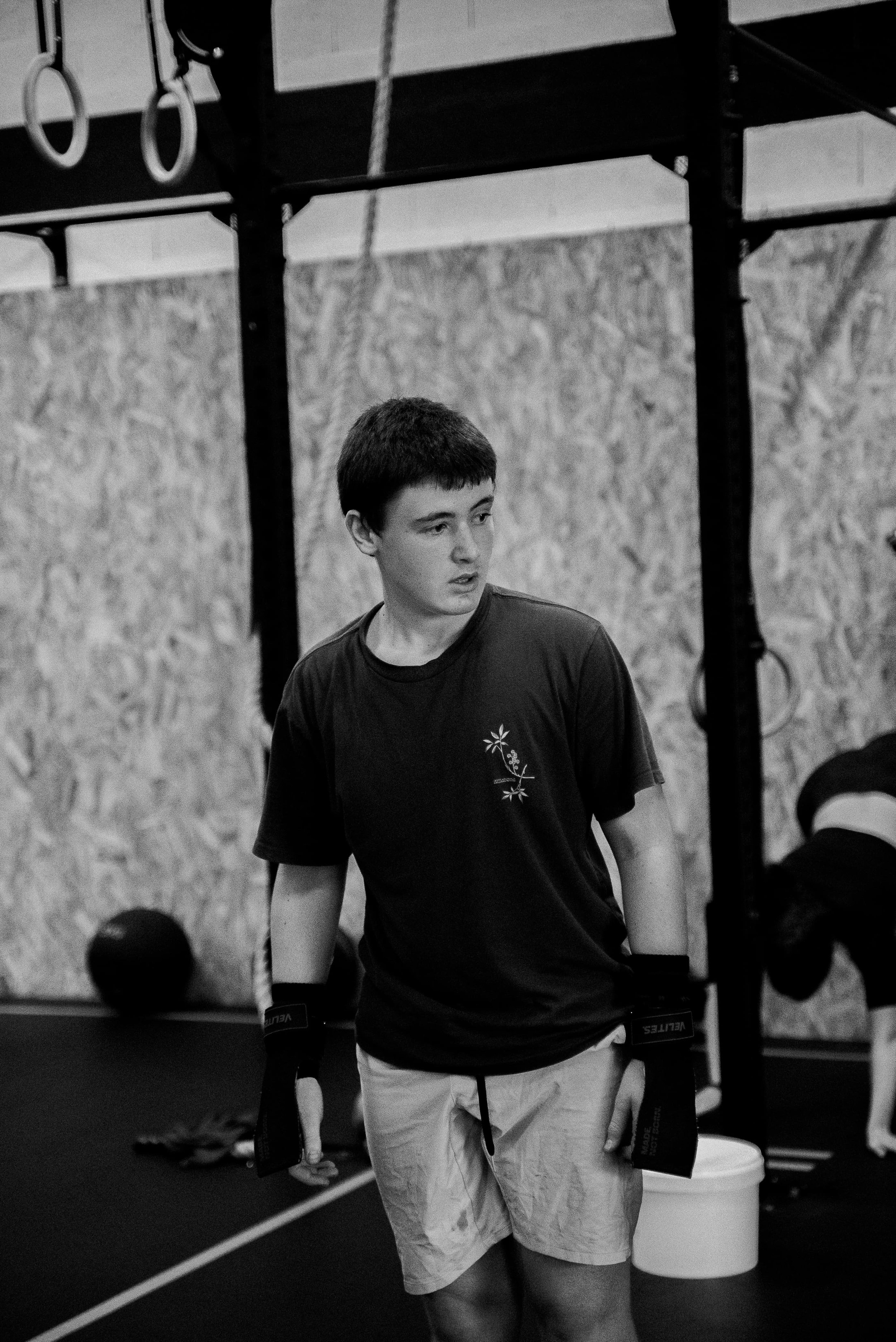 Jeune homme en t-shirt noir dans un gymnase, entouré d'équipement sportif et d'anneaux de gymnastique.