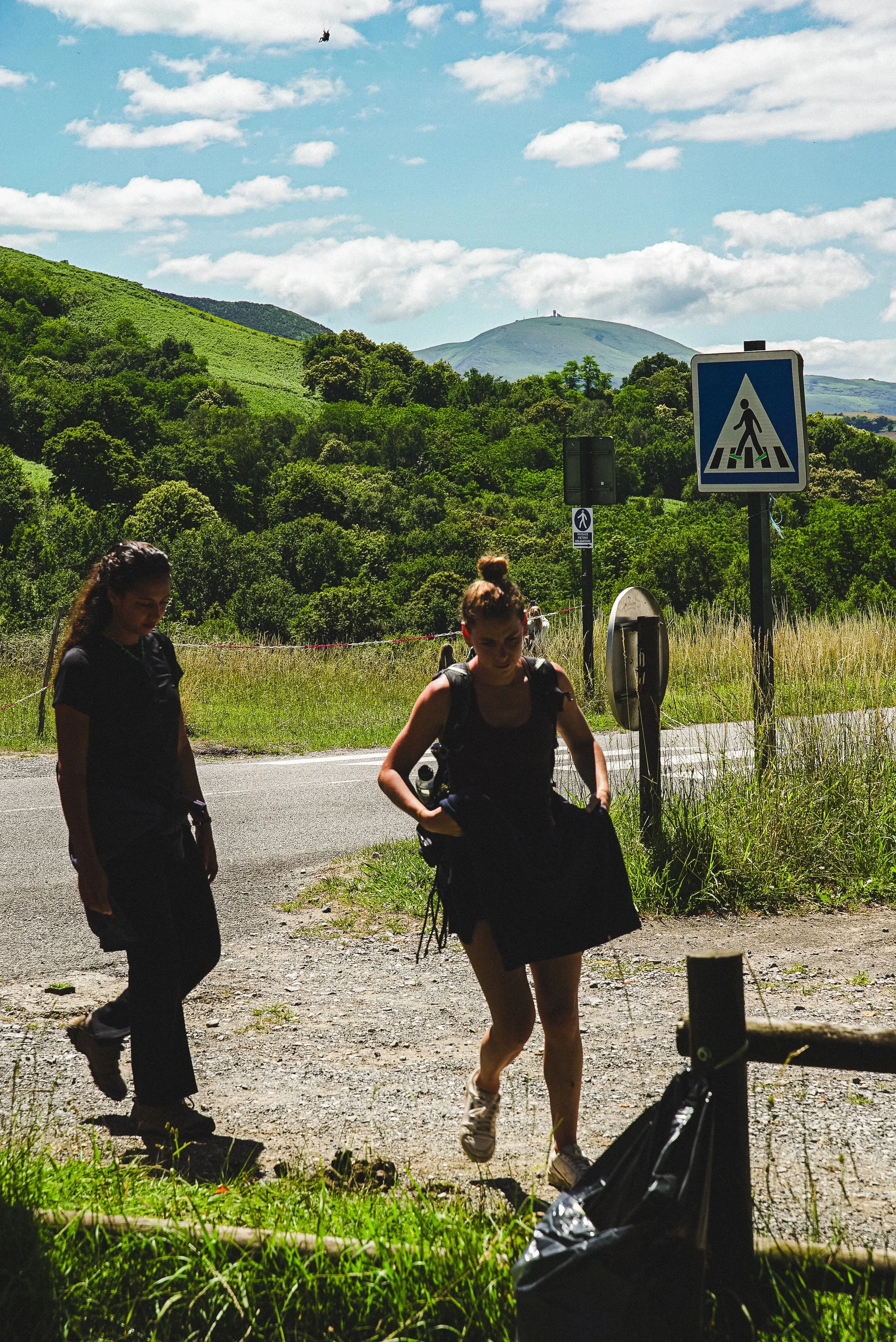 Trois femmes portant des sacs à dos marchent sur le bord de la route en pleine campagne avec des collines verdoyantes en arrière-plan, un panneau de passage piéton visible, un paysage ensoleillé avec un ciel partiellement nuageux et une personne fais