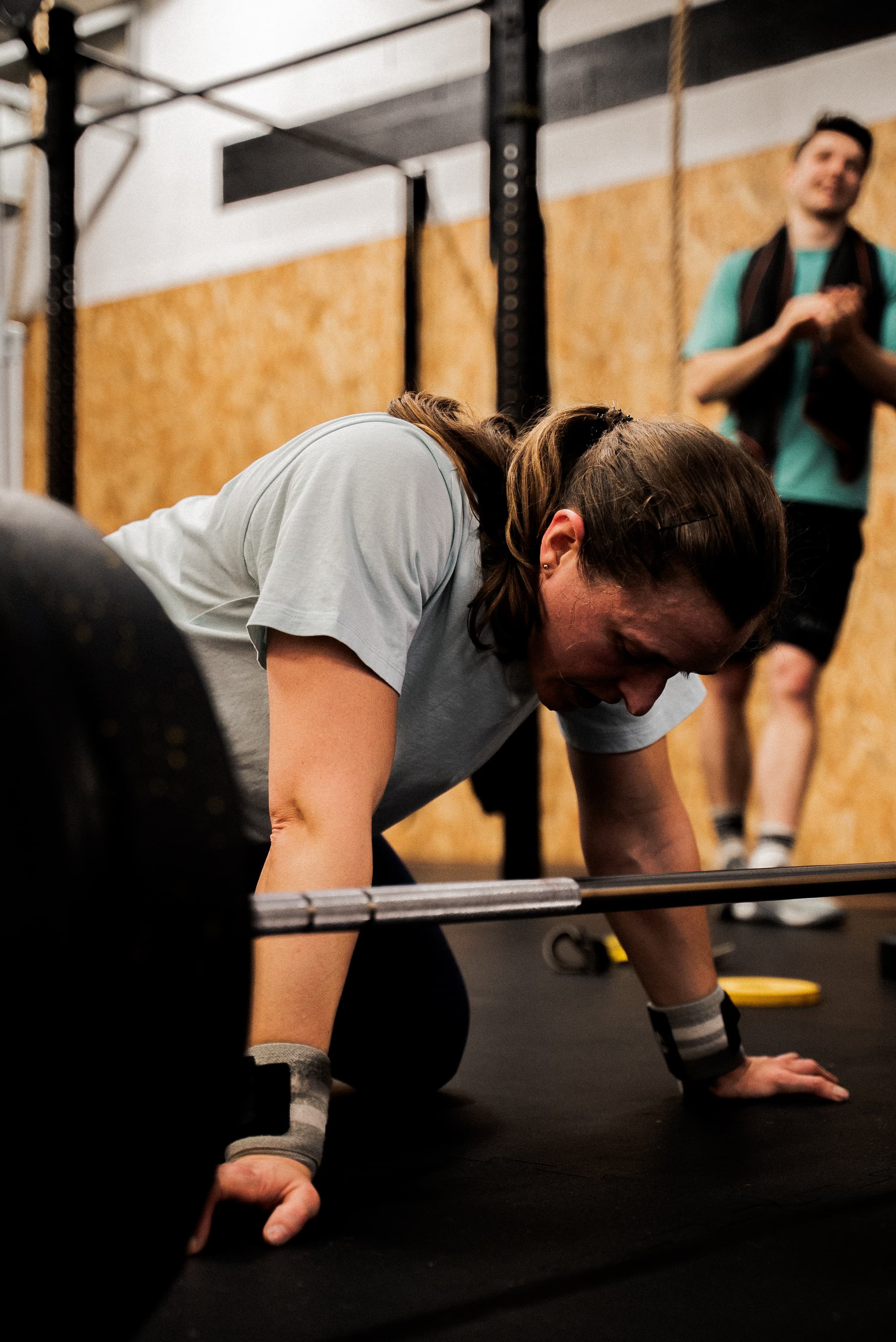 Une femme effectuant un exercice de musculation au sol dans une salle de sport, avec un homme en arrière-plan qui regarde et sourit.