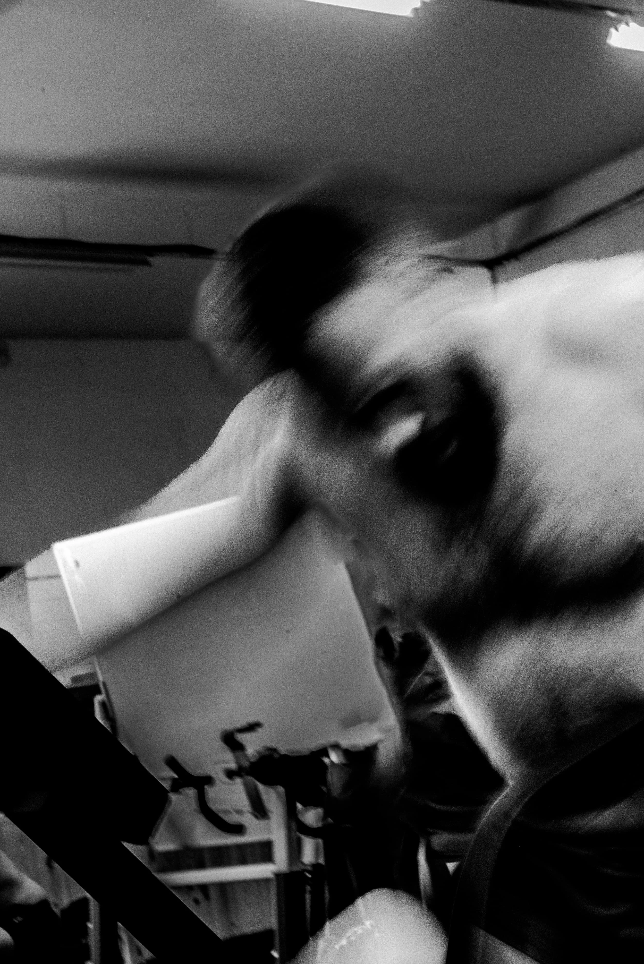 Photo en noir et blanc d'un chat noir et blanc assis sur une table ou un comptoir, à côté d'une tasse ou un récipient. En arrière-plan, il y a des meubles ou étagères.