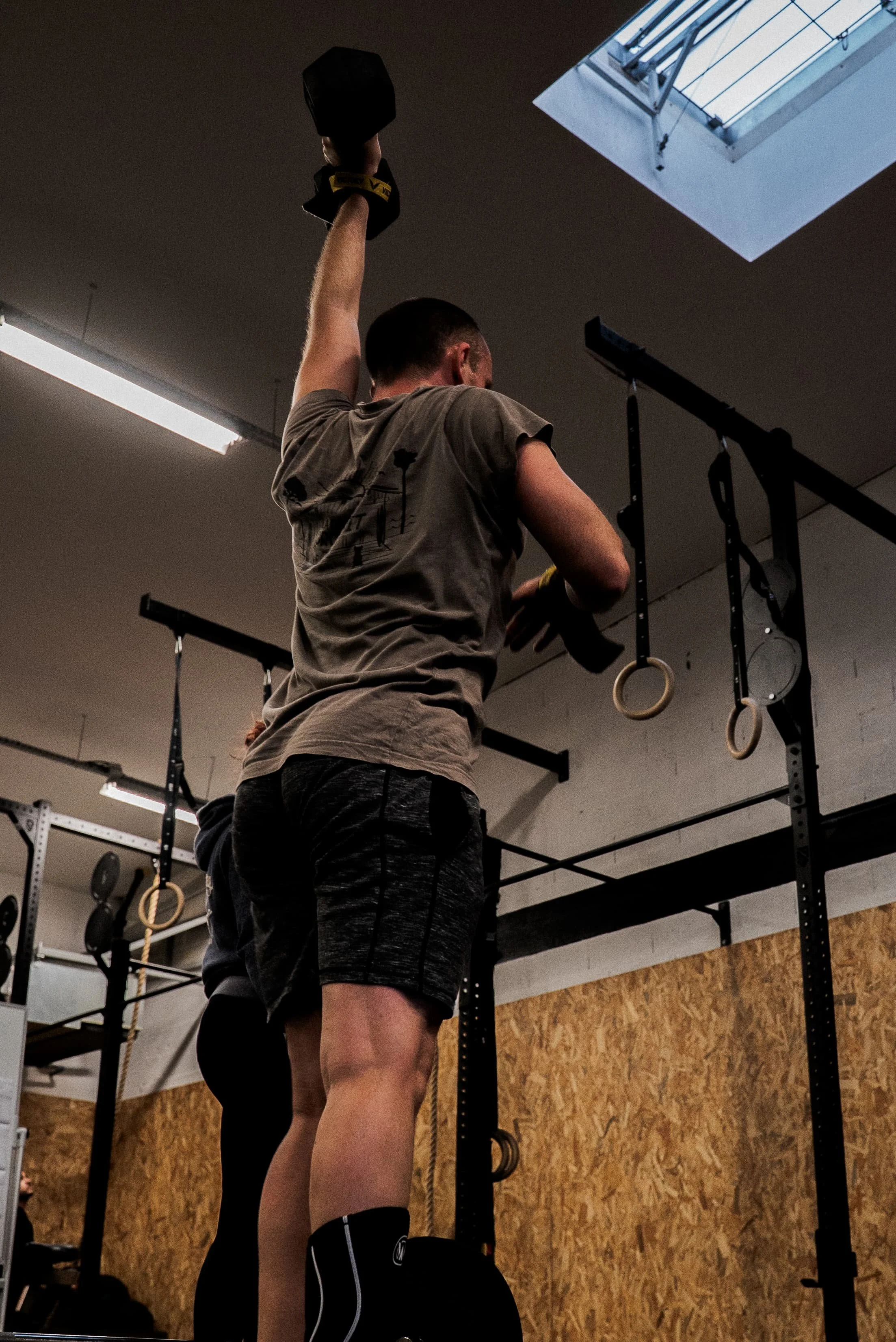 Un homme en t-shirt gris et short noir effectue un entraînement de musculation avec un haltère dans une salle de sport, à côté d'une structure avec des anneaux de gymnastique suspendus.