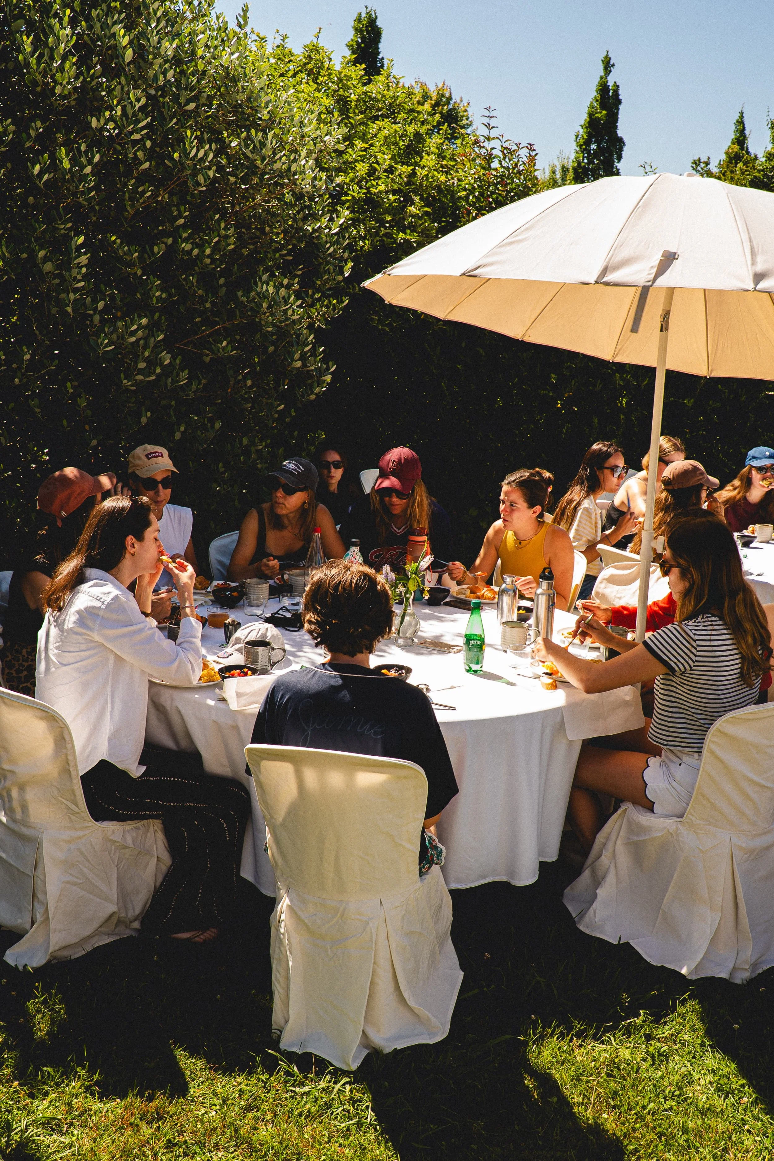 Groupe de personnes assises autour d'une table blanche en plein air sous un parasol beige, qui prennent un repas ensoleillé dans un jardin avec des arbustes et des arbres en arrière-plan.