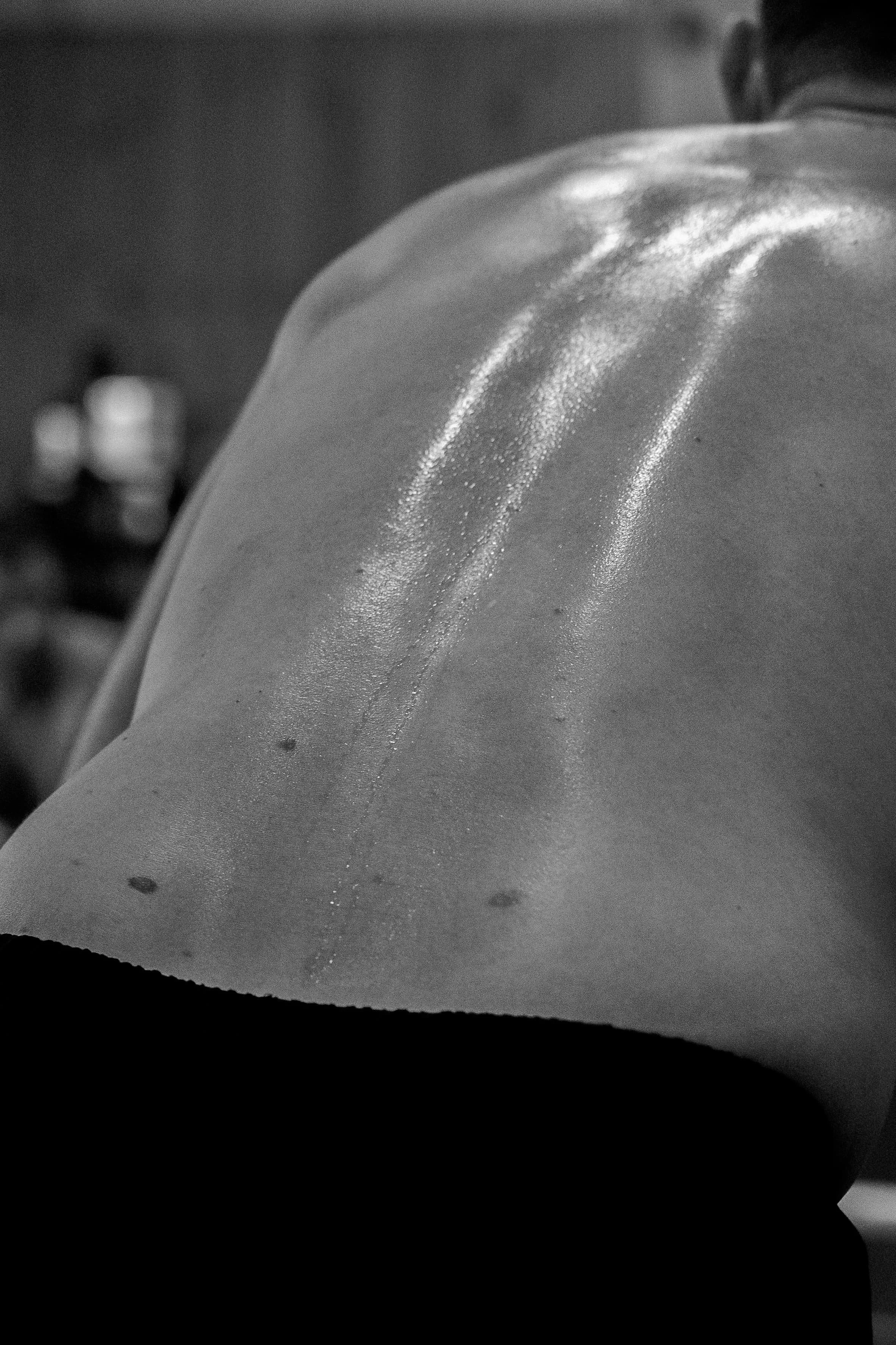 Photo en noir et blanc d'une personne avec des épaules nues, vue de dos, avec une lumière refletant sur sa peau, mettant en valeur la texture de sa peau.