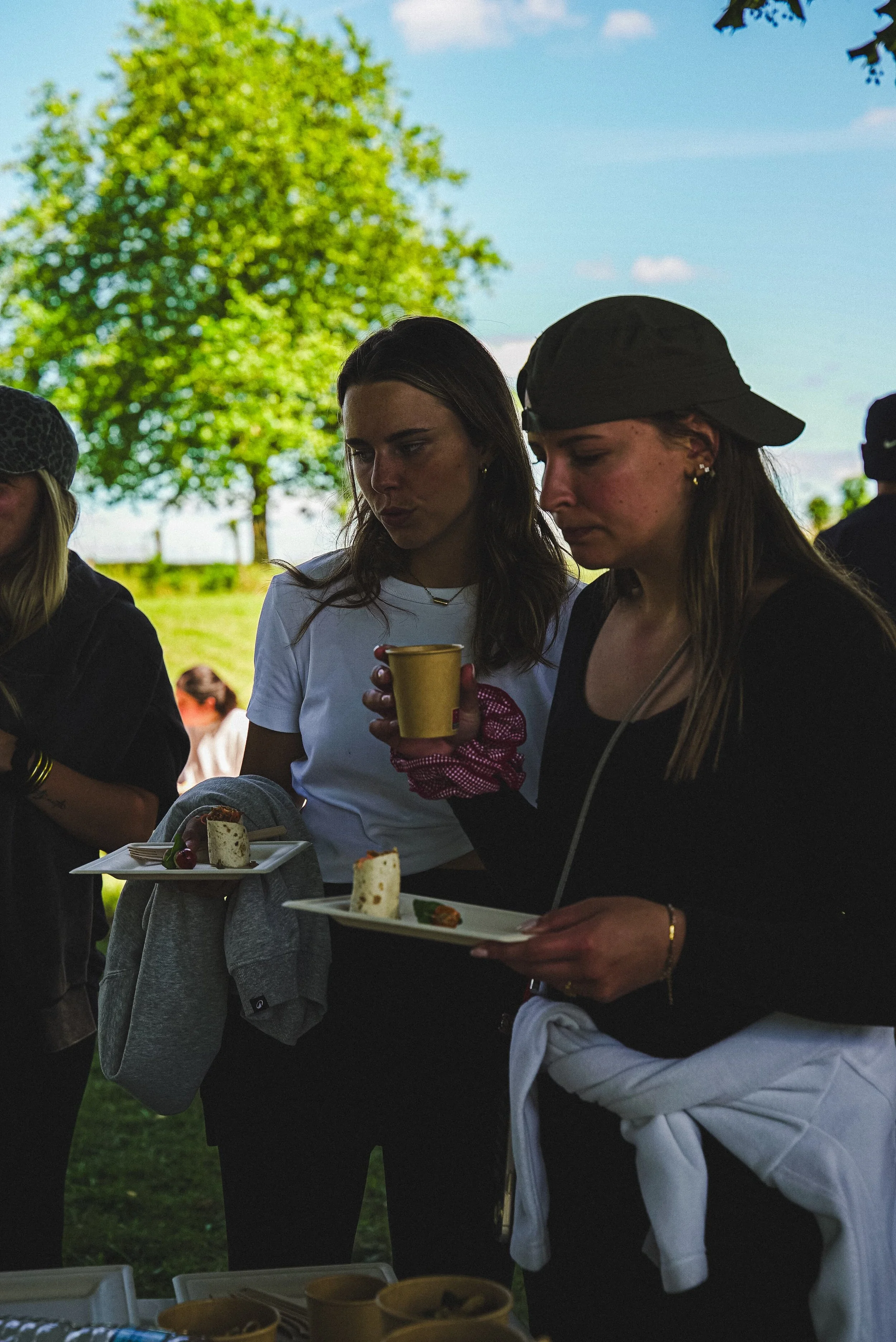 Deux femmes examineant des plats de nourriture lors d'un événement en plein air, avec un arbre en arrière-plan.