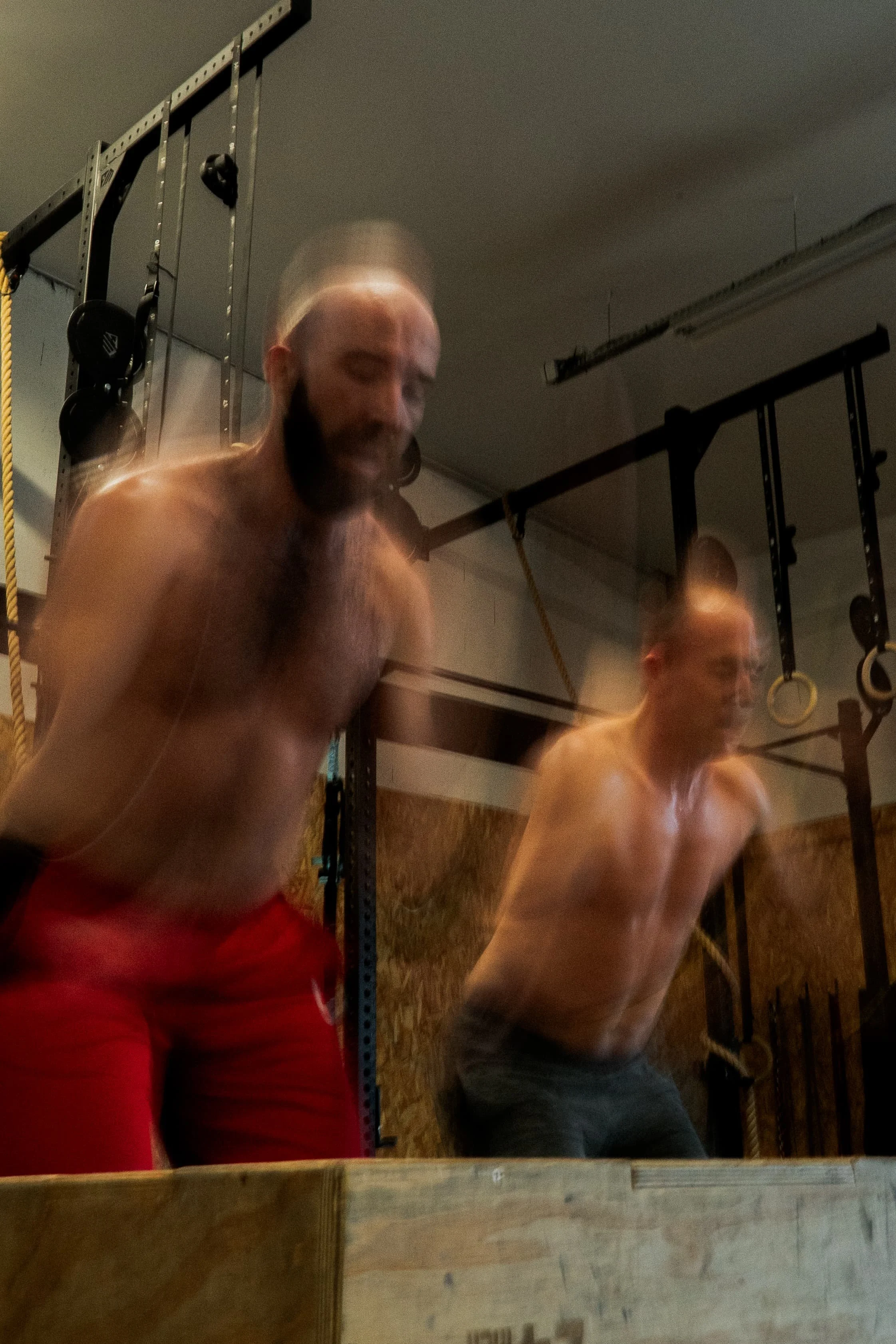 Deux hommes nus à l'intérieur d'une salle de sport, probablement en train de s'entraîner ou de se préparer pour un exercice, avec du matériel de sport en arrière-plan.