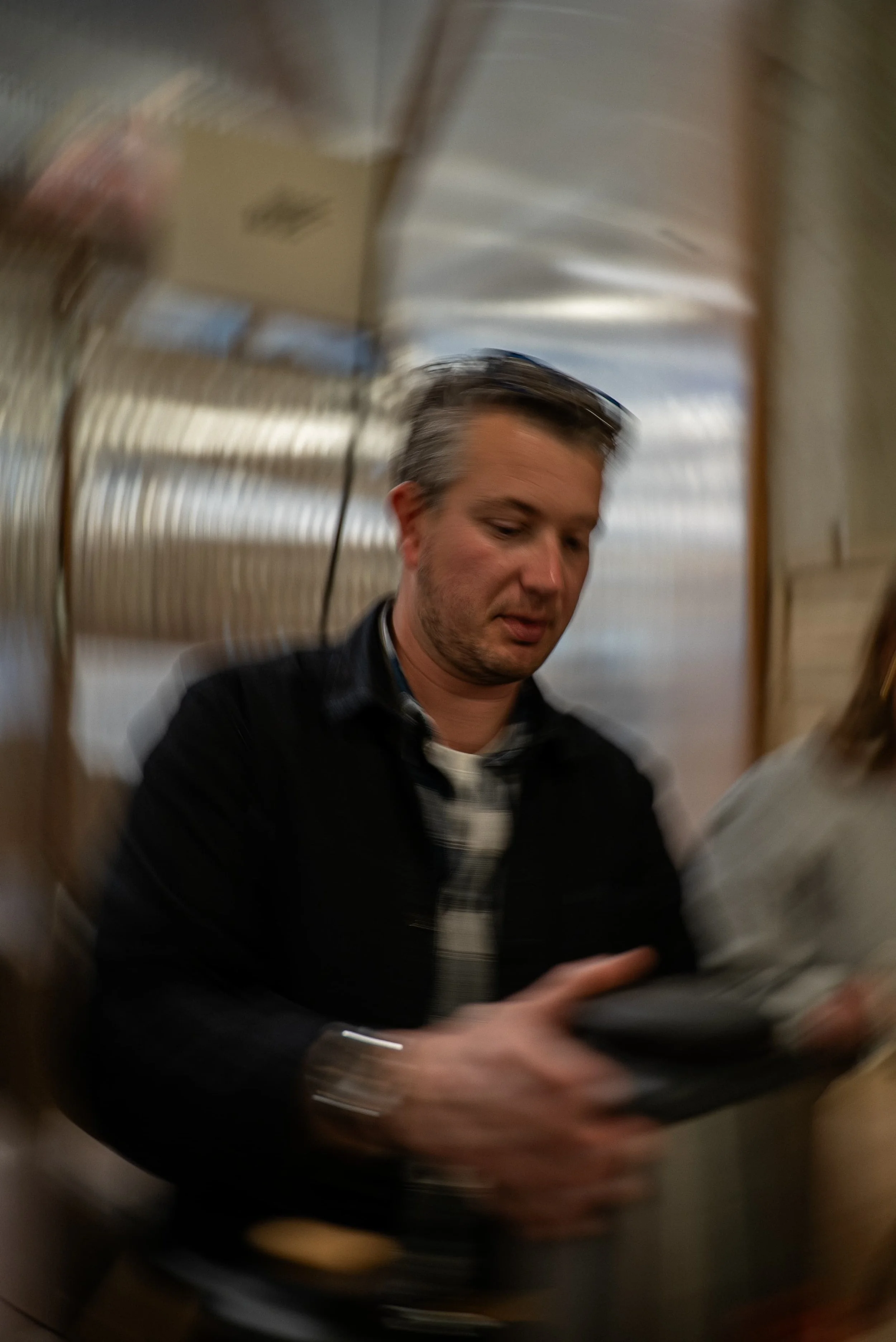 Portrait flou d'un homme avec des cheveux gris, portant un pull noir avec une chemise à motifs noir et blanc, étant dans un environnement métallique ou en tunnel.