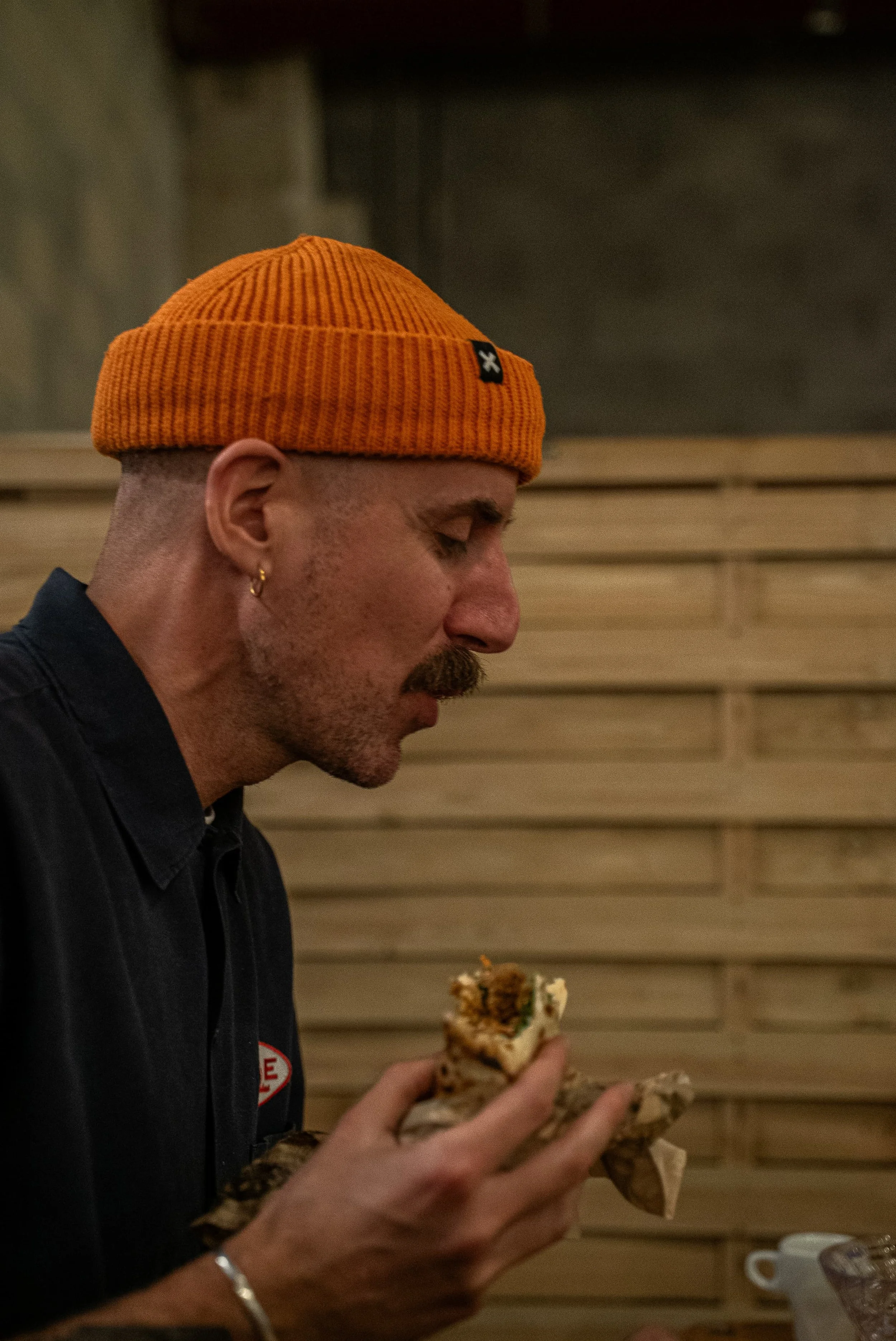 Un homme portant un bonnet orange et une veste noire tient un morceau de gâteau en main lors d'un repas dans un environnement intérieur.