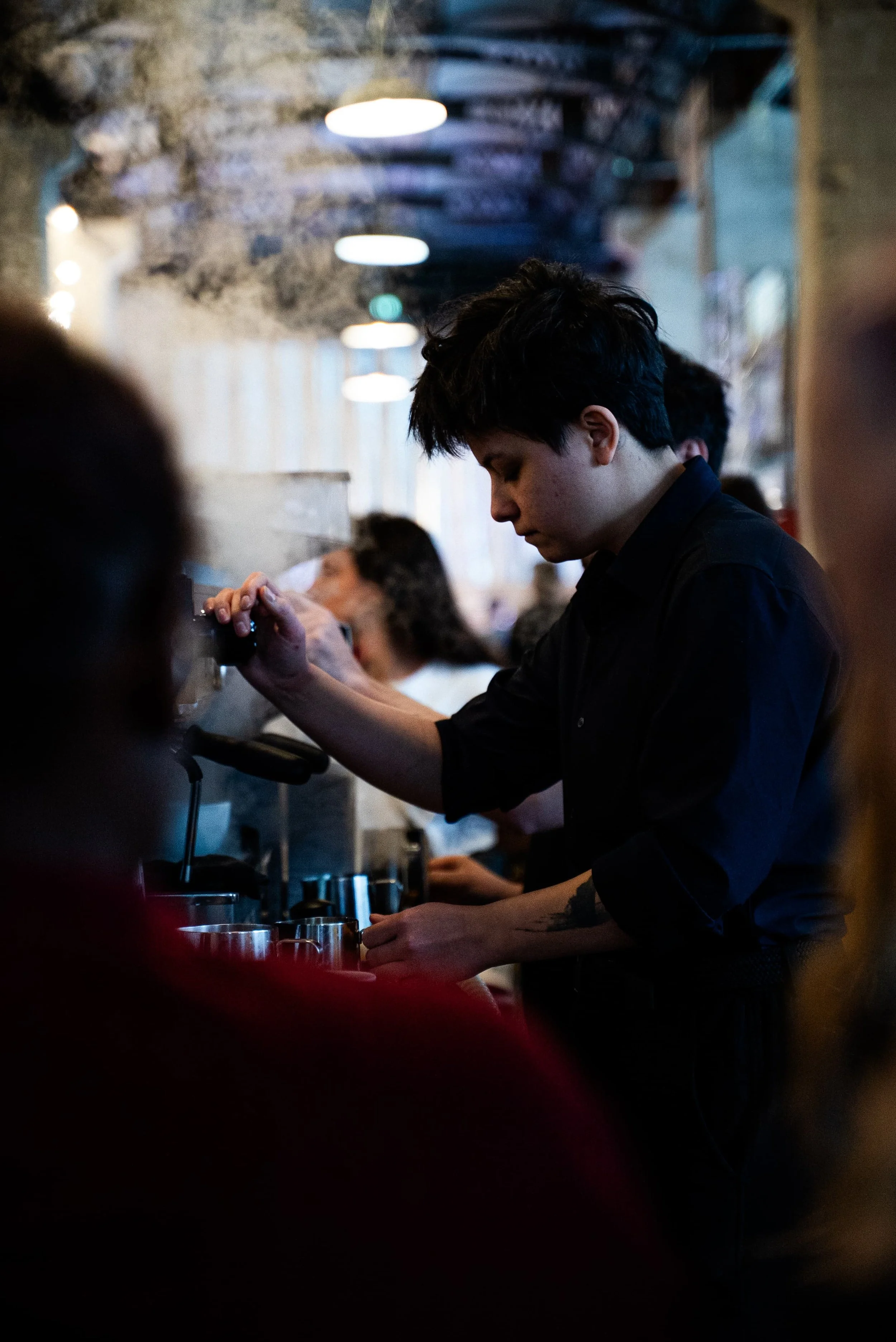 Un jeune homme au visage concentré, face à un comptoir de café ou de bar, en arrière-plan, une femme semble préparer une boisson, dans un intérieur à l'ambiance chaleureuse et intime.