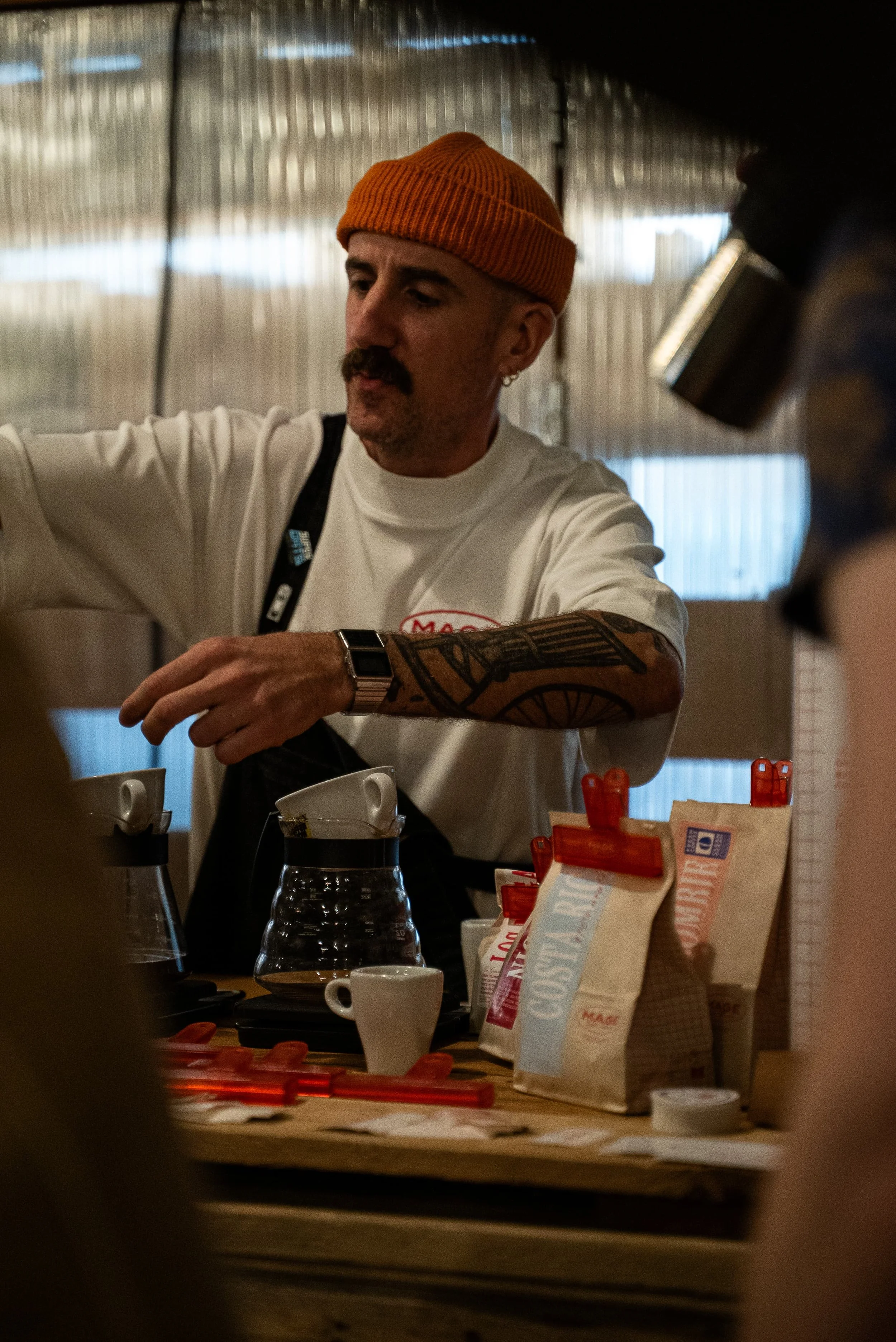 Un homme portant un bonnet orange, une chemise blanche et une montre regarde à gauche. Il se trouve dans un intérieur, entouré de sacs de café et de tasses, avec un fond en bois et un mur en panneaux de plastique transparent.
