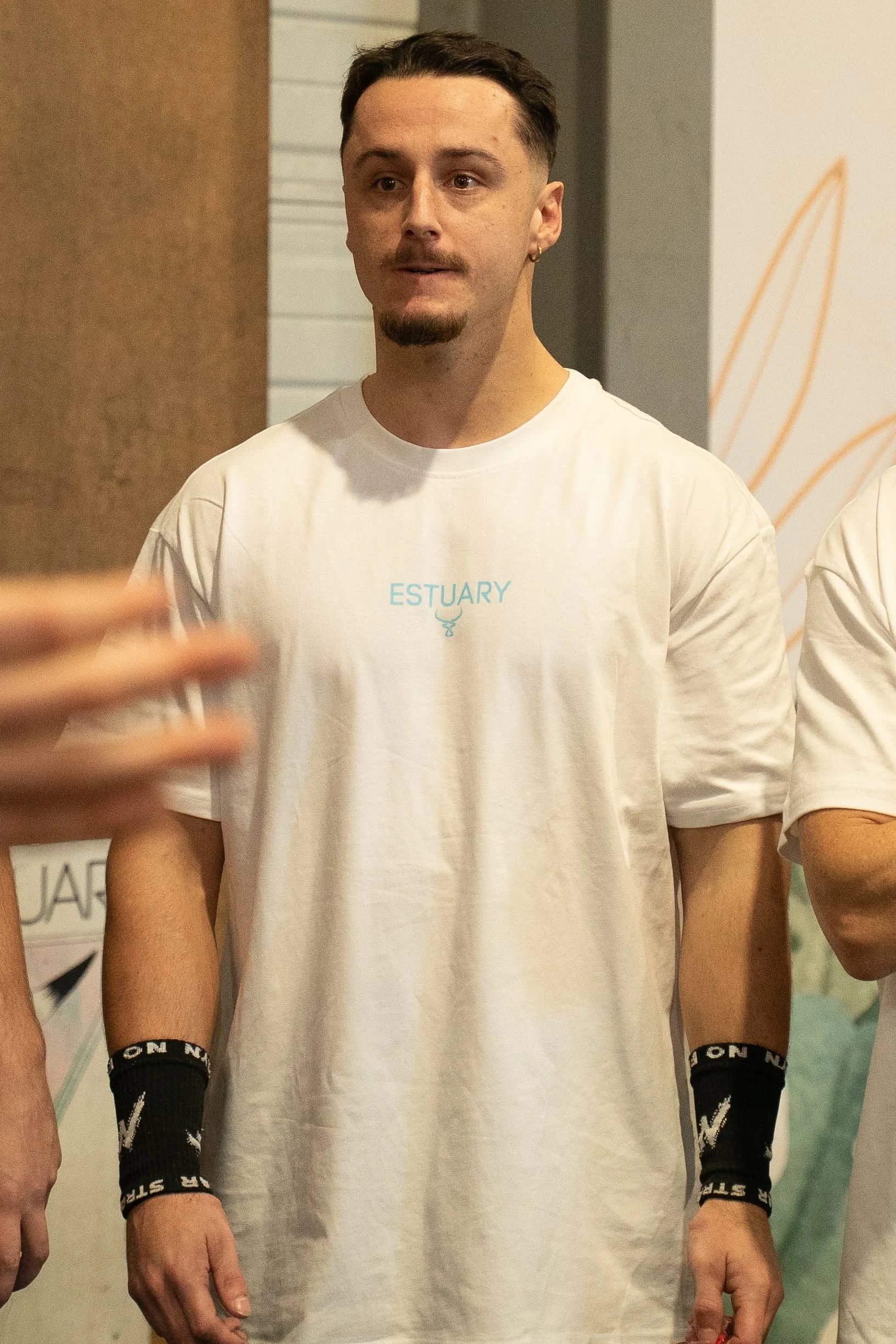 Image d'un homme portant un t-shirt blanc avec le mot 'ESTUARY' et une illustration de narval, en train de se faire prendre en photo dans un endroit intérieur.