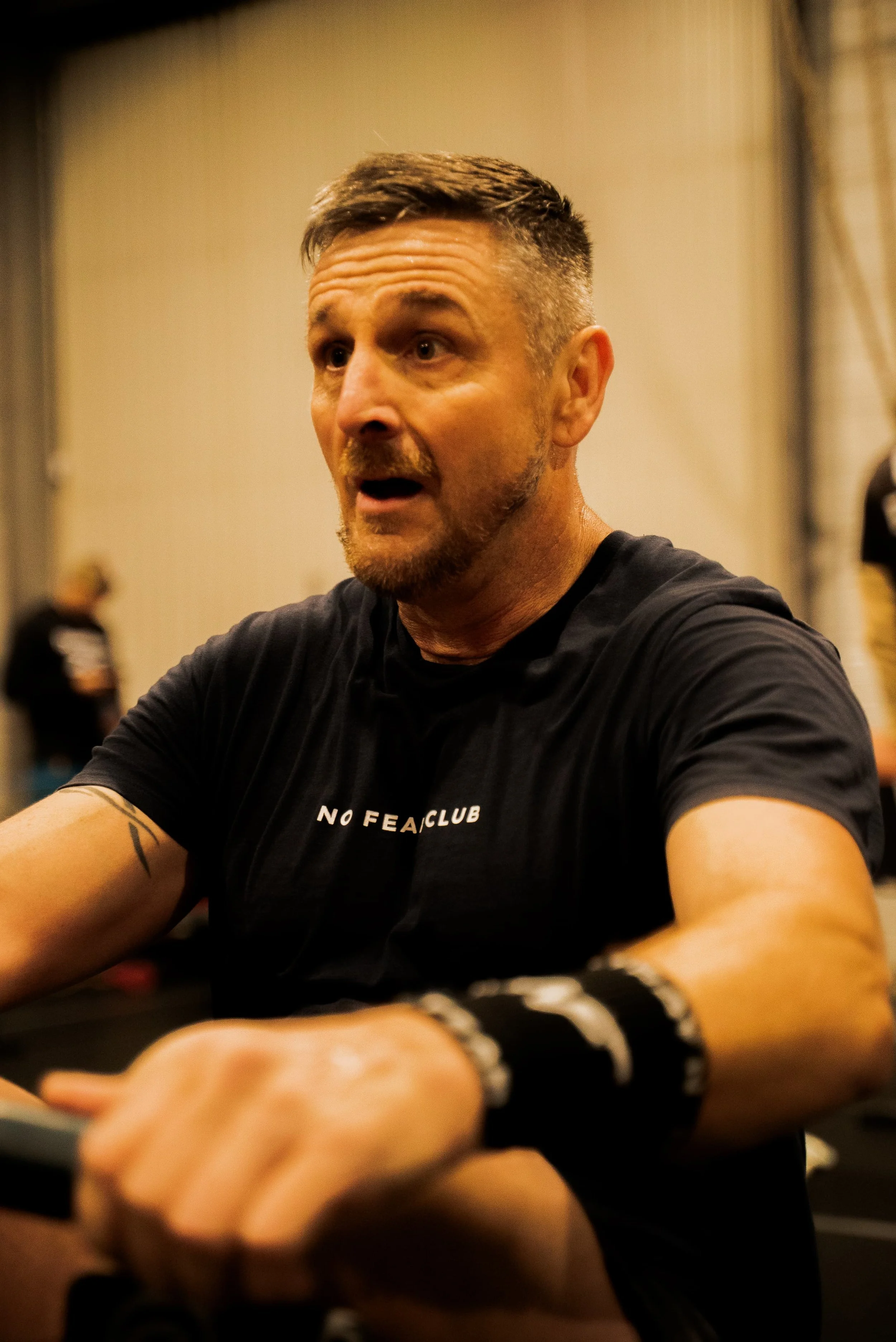 Homme aux cheveux courts et barbe, portant un t-shirt noir avec inscription "NO FEAR CLUB", en train de prendre un selfie dans ce qui semble être une salle de sport.