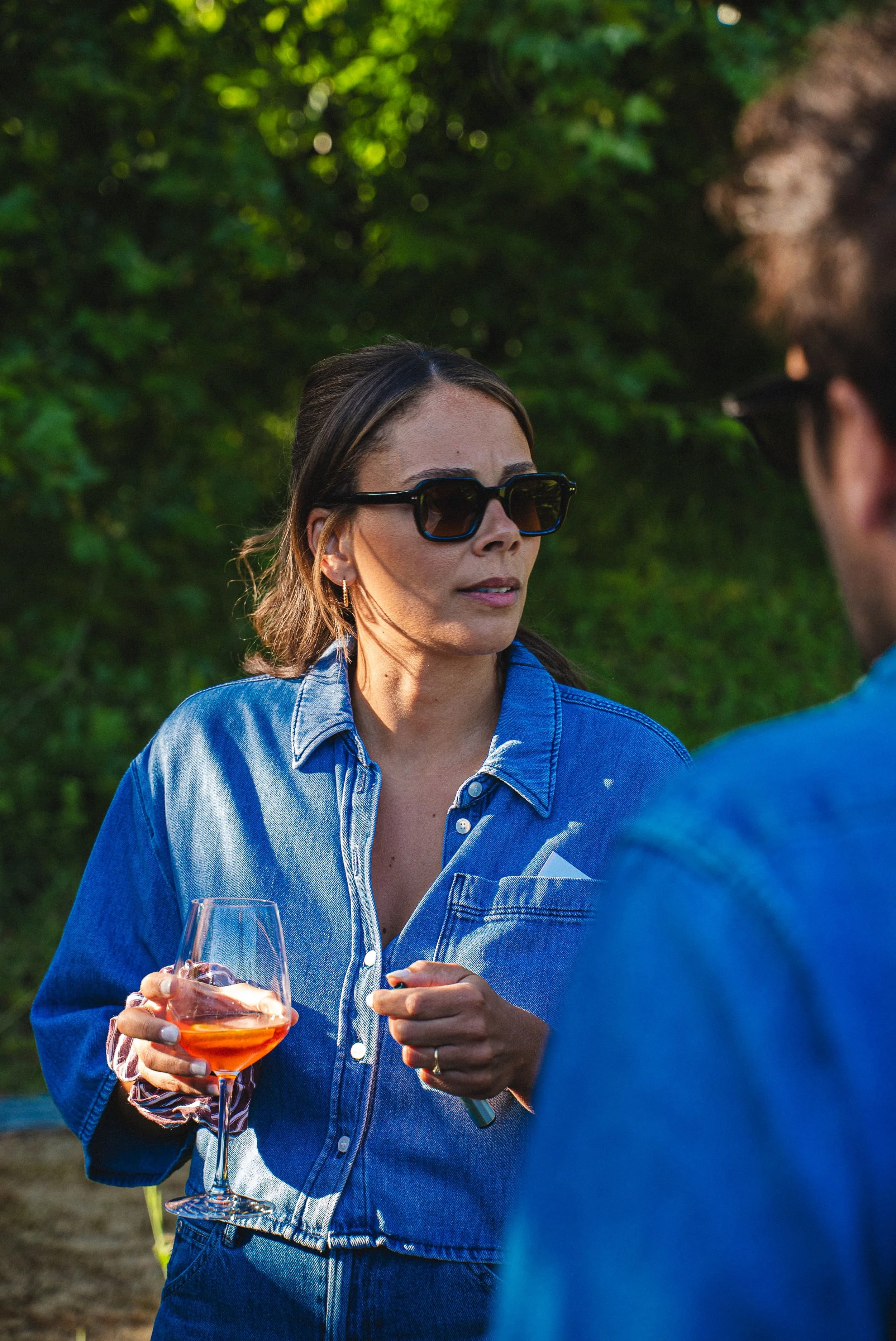 Une femme avec des lunettes de soleil, portant une chemise en jean, tenant un verre de vin rosé, en conversation avec un homme dans un cadre extérieur avec une végétation verte en arrière-plan.