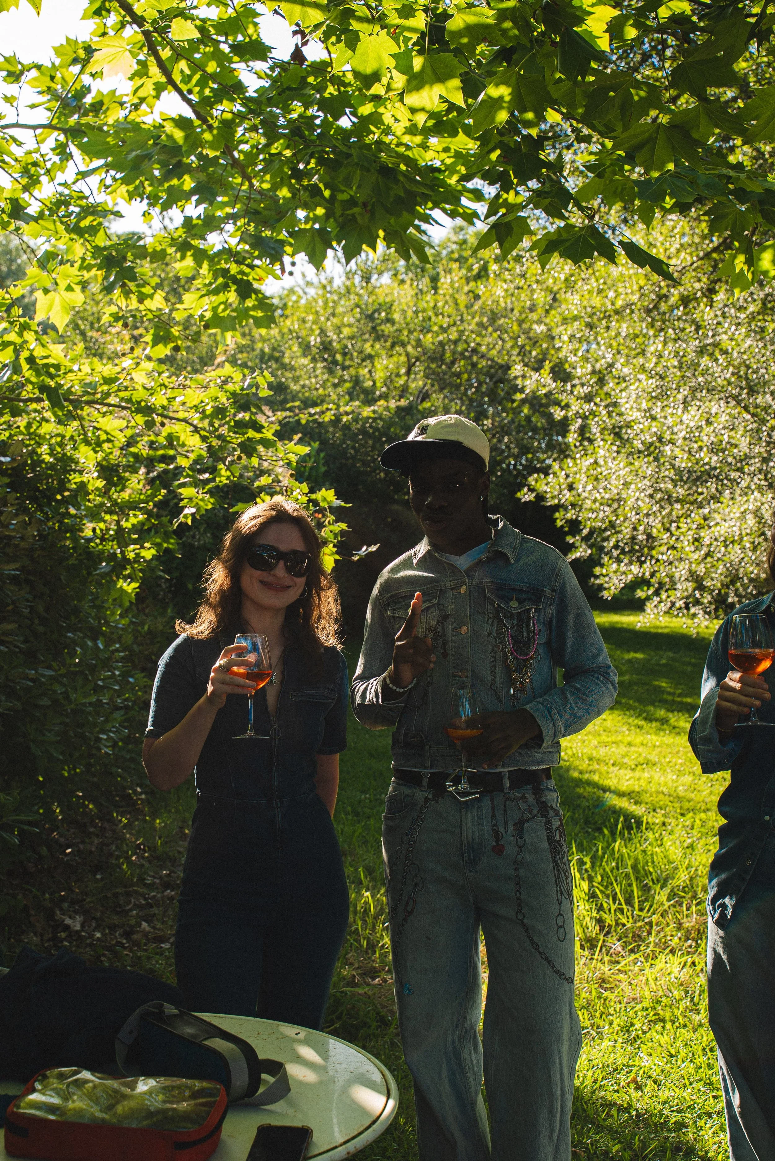 Des personnes qui tiennent des verres de vin rosé en extérieur sous un arbre ensoleillé, avec un groupe de personnes visibles en arrière-plan.