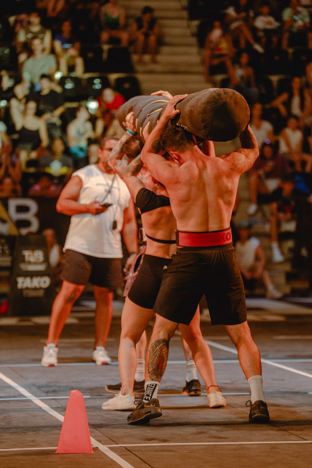 Deux athlètes musclés soulevant un sac lourd lors d'une compétition de fitness avec public dans les gradins — photographe fitness Voar by Tim