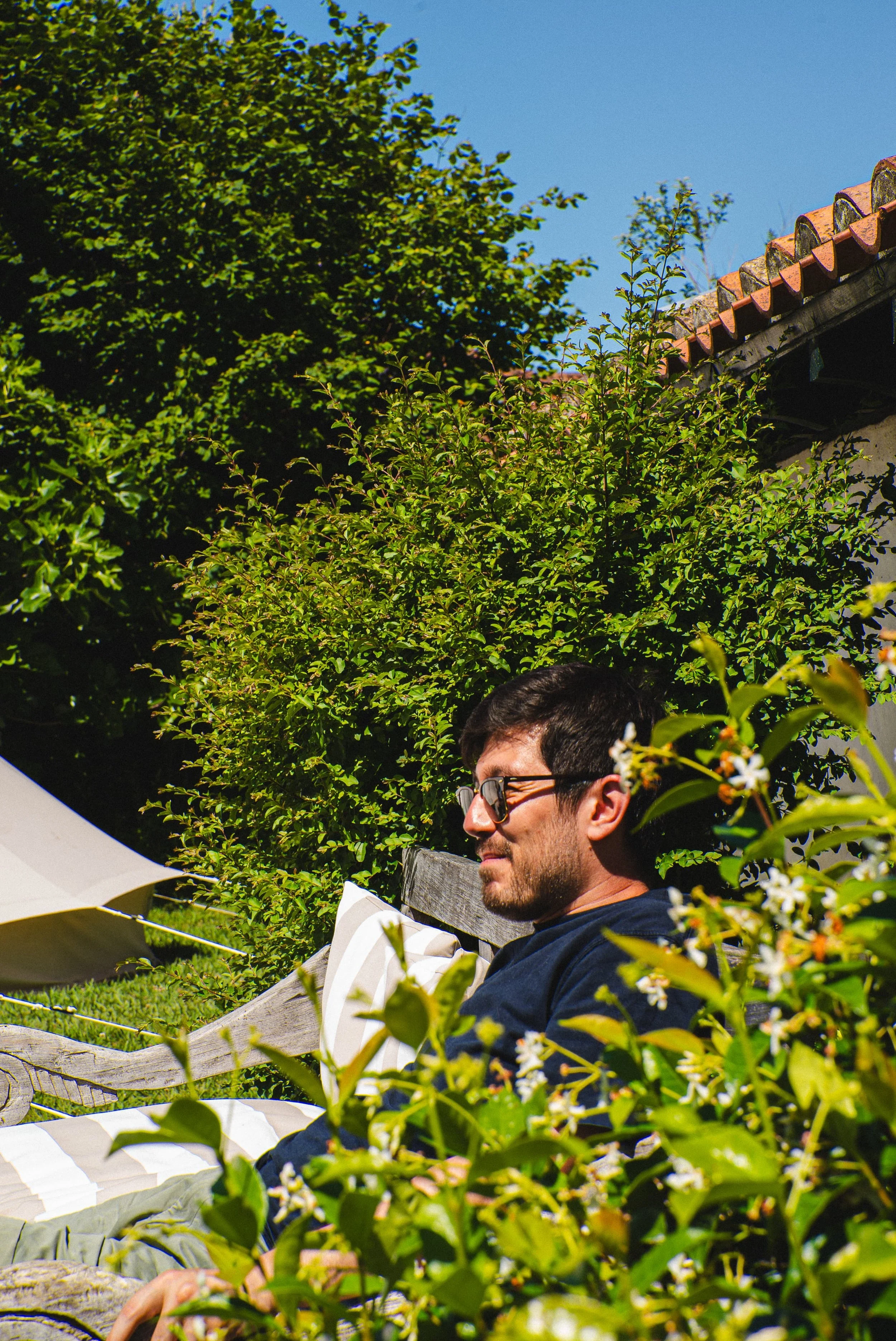 Un homme avec des lunettes assis dans un jardin, entouré de verdure et sous un ciel bleu, en train de lire ou de se détendre.