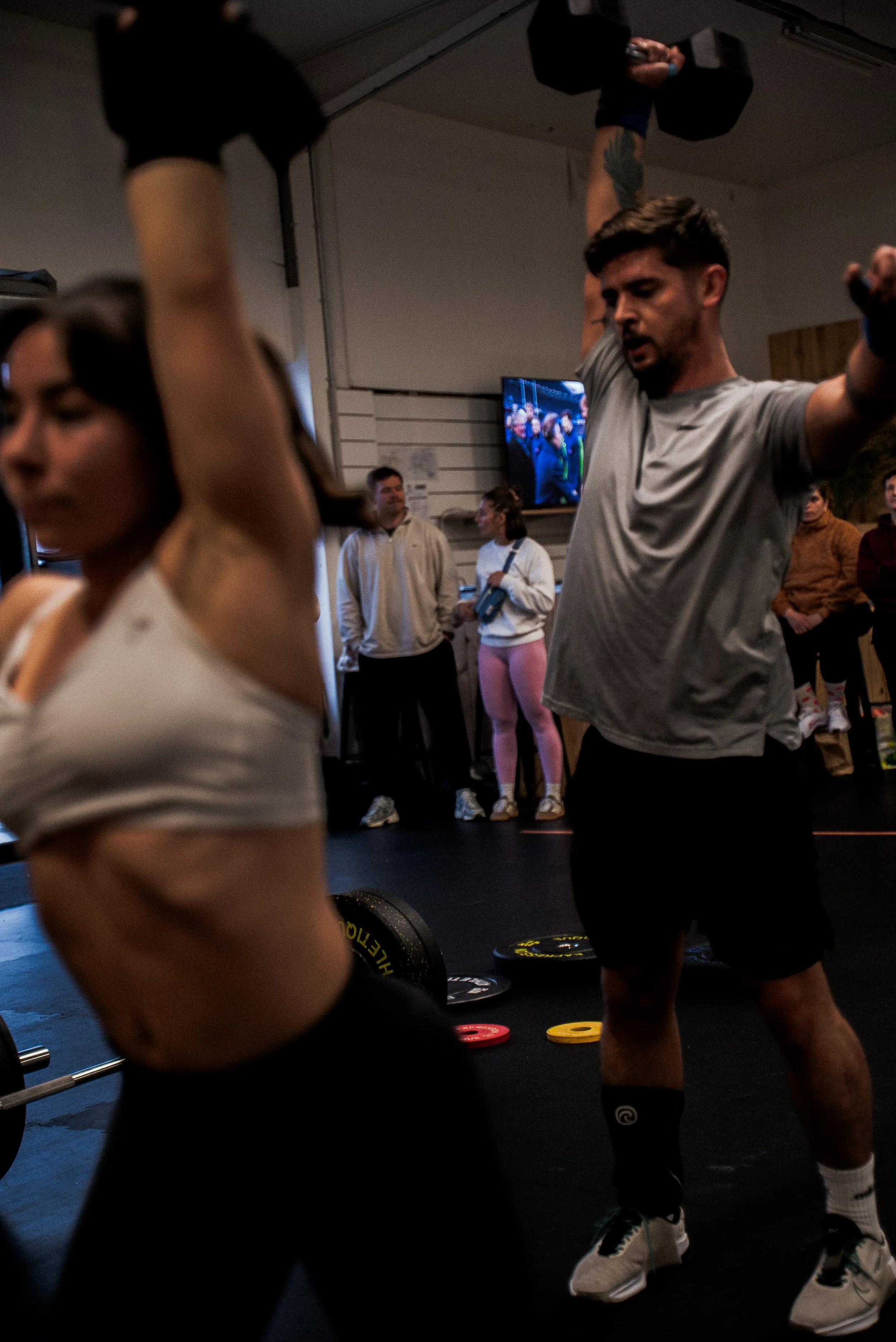 Une personne en train de faire un entraînement de musculation avec des haltères dans une salle de sport, entourée d'autres personnes qui regardent.