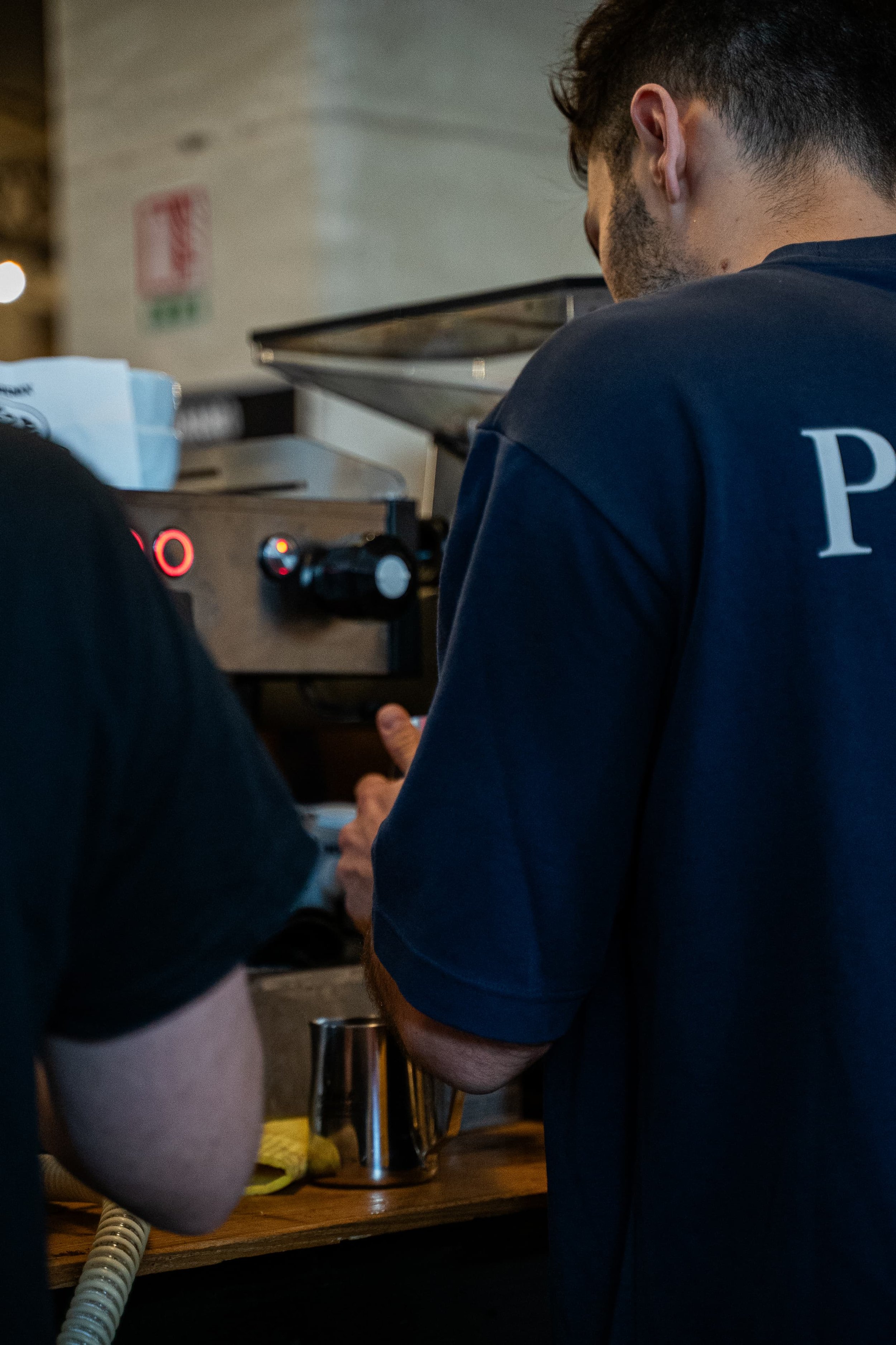 Un homme vêtu d'un t-shirt bleu avec une lettre blanche, vu de dos, en train de travailler ou de préparer quelque chose derrière une surface en bois, avec un espace de cuisine ou de travail visible en arrière-plan.