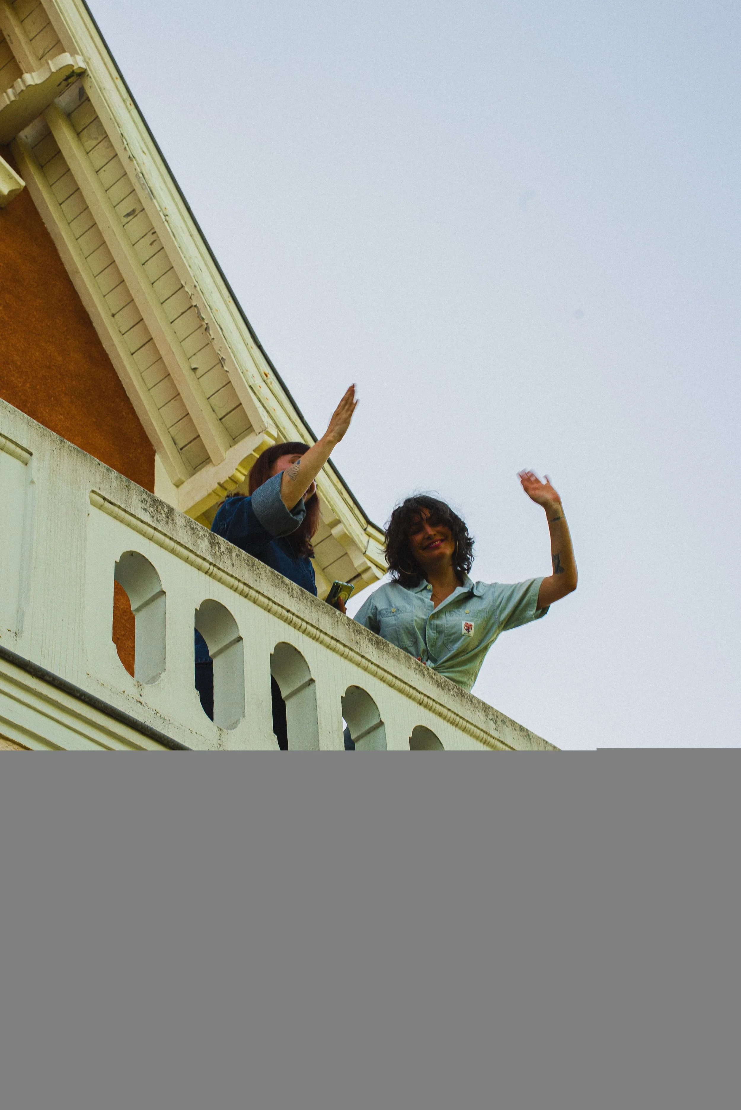 Deux femmes souriantes saluent depuis un balcon en haut d'un bâtiment, saluant le public lors d'une journée ensoleillée.