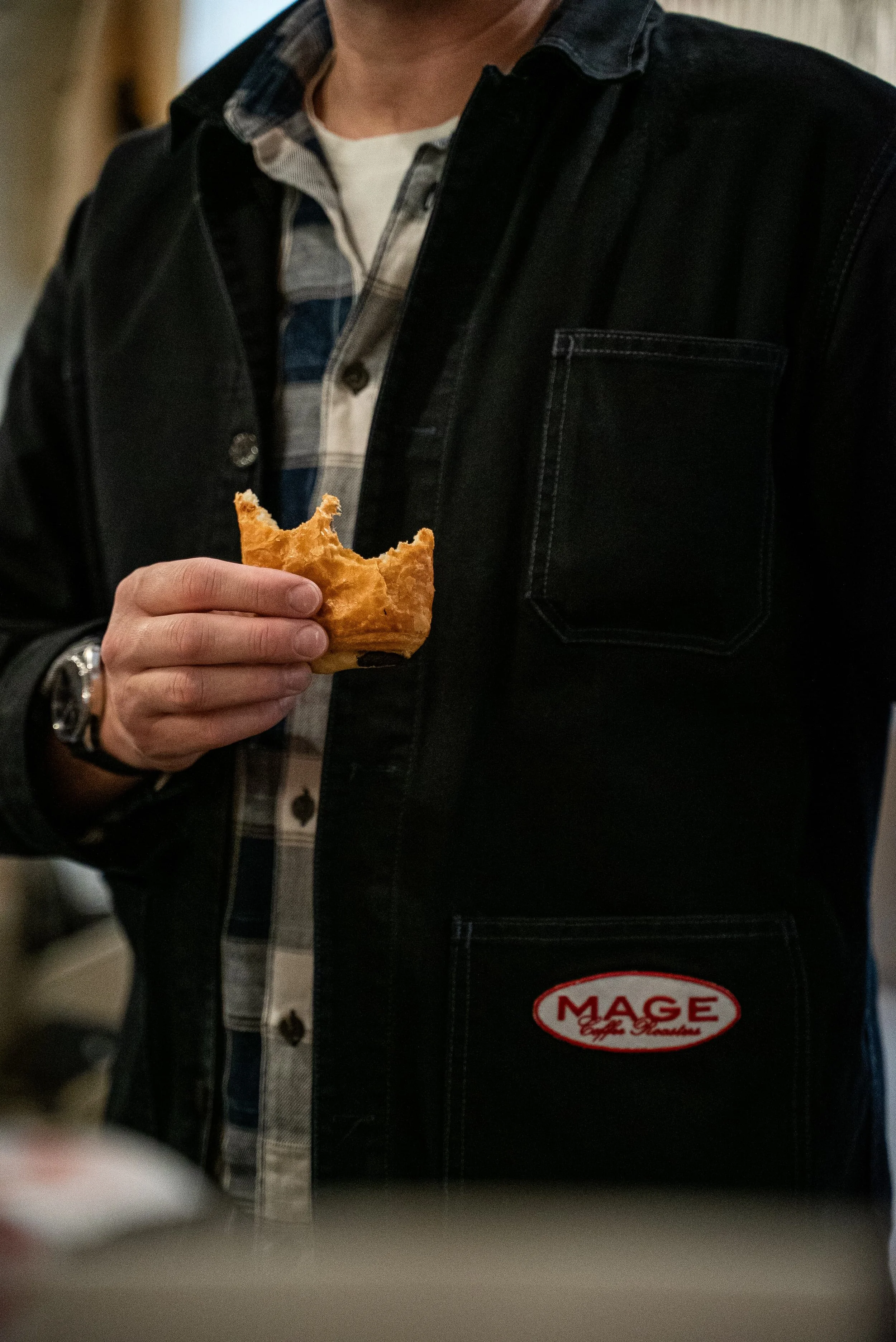 Personne tenant un croissant dans une main, portant une veste noire avec un badge "MAGE" sur la poitrine, dans un environnement intérieur.