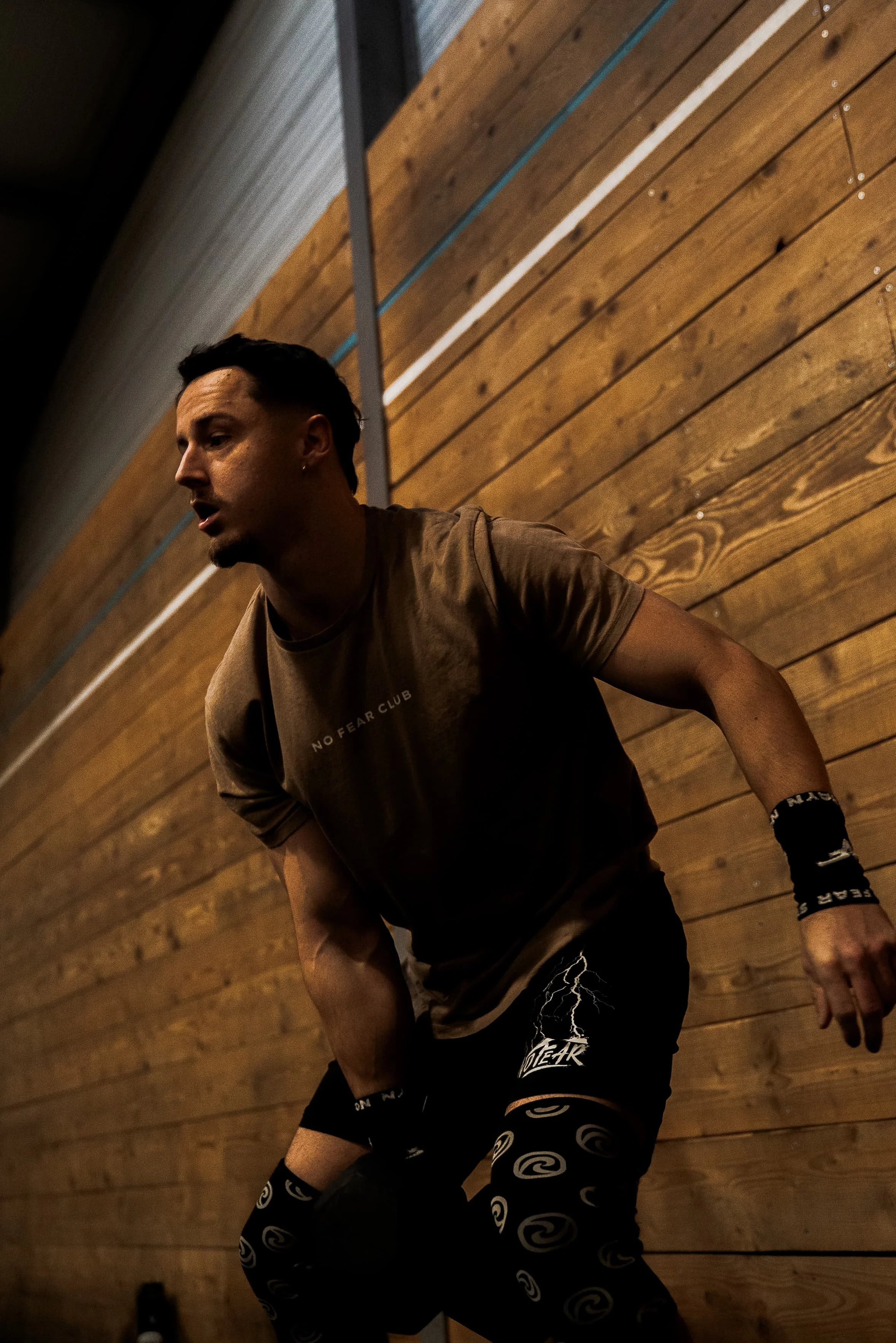 Un homme en vêtements de sport en position de squat dans une salle avec un mur en bois derrière lui.