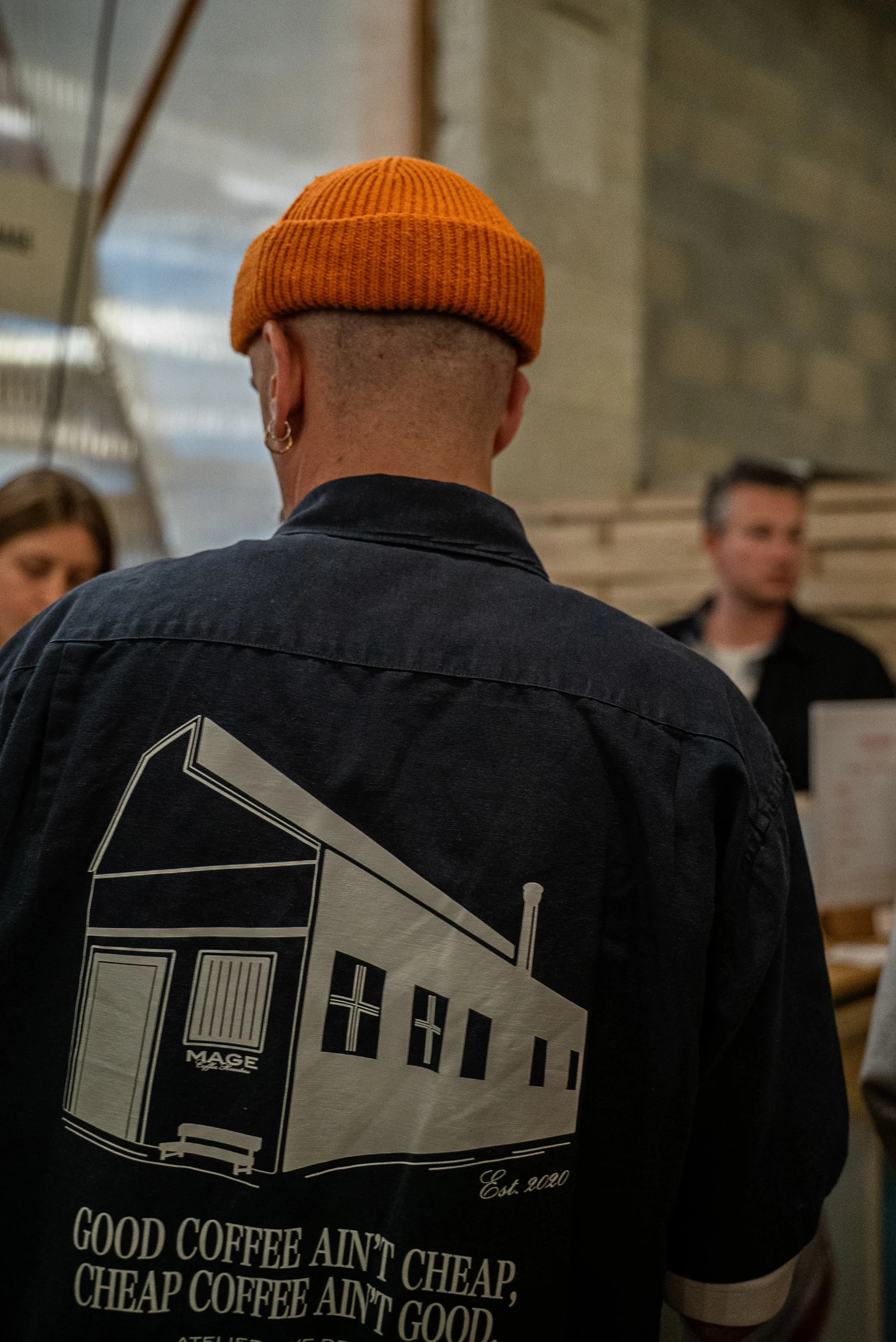 Photo de dos d'un homme portant un bonnet orange et une veste noire avec un graphisme blanc à l'arrière, dans un lieu intérieur, avec d'autres personnes en arrière-plan.