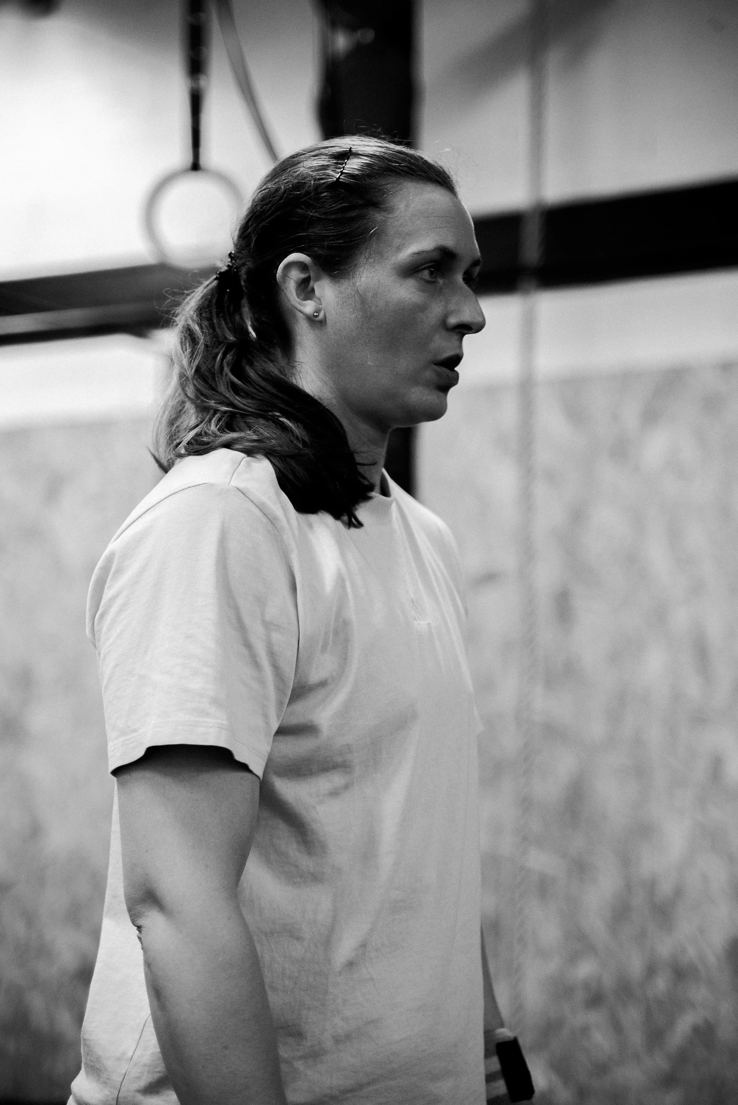 Une femme en profil, portant un t-shirt, dans un environnement intérieur avec des murs en briques et équipement de gymnastique, comme des anneaux de gymnastique suspendus.