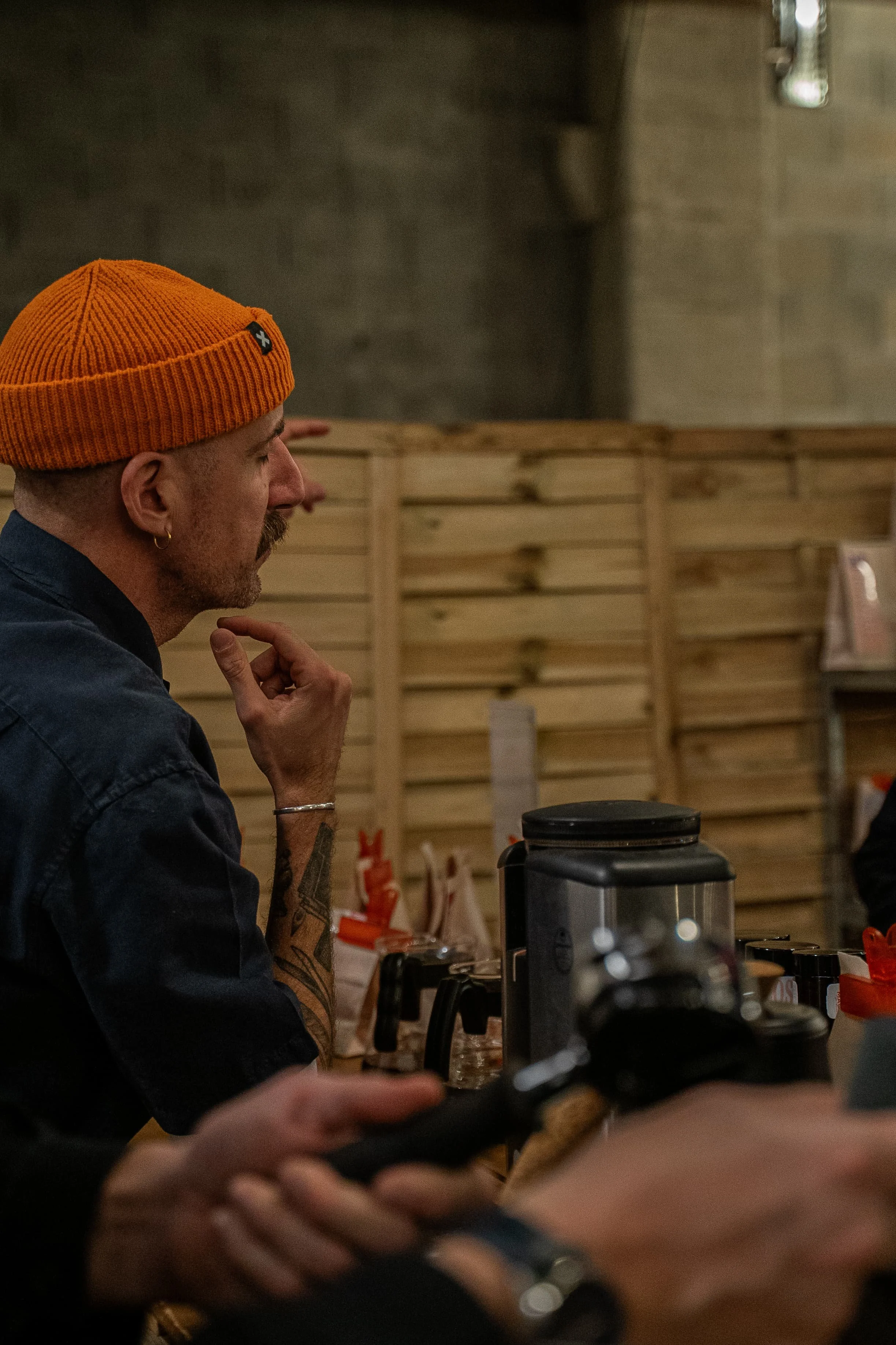 Un homme portant un bonnet orange et une boucle d'oreille en or est assis dans un environnement intérieur avec un fond en bois et un mur en béton. Il semble être en train de regarder quelque chose, peut-être un appareil de café.