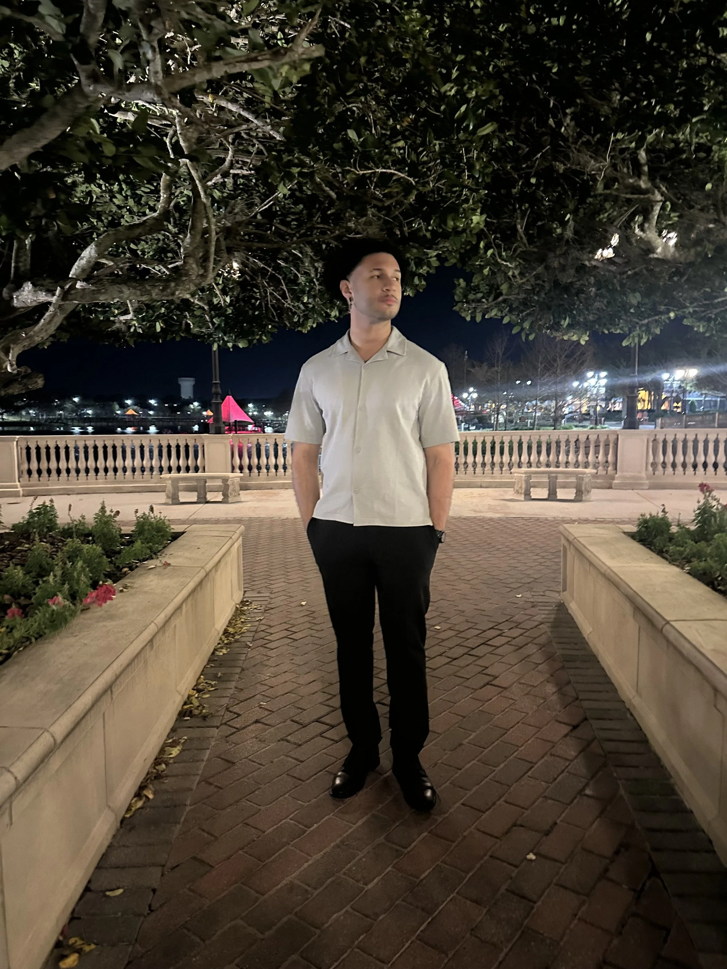 A young man in a beige short-sleeved button-up shirt and black pants stands outdoors at night under a large tree. The background features a lit waterfront area with colorful lights and structures, and a railing along a walkway. The man has his hands in his pockets and is looking slightly to his right with a neutral expression.