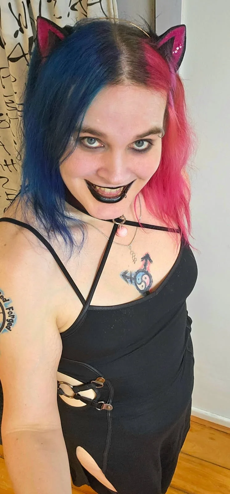 Woman with half blue and half pink hair wearing cat ears headband, black dress with cutouts, and black lipstick, smiling at the camera.