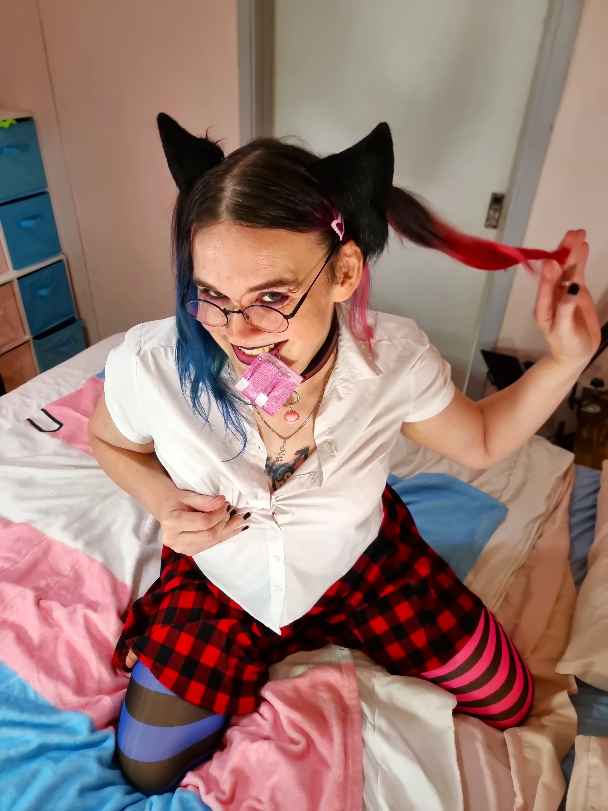A Rockhampton Transgender woman sex worker wearing glasses cat ears and has her hair up in pigtails. She is smiling suggestively and has a condom in the wrapper hanging from between her teeth.